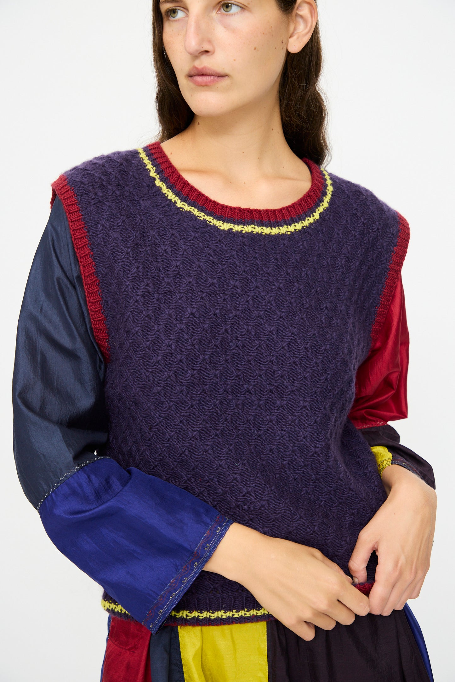 A person wears the Injiri Merino Wool Sweater Vest in Purple, Red, and Yellow over a long-sleeved shirt, standing against a plain background.