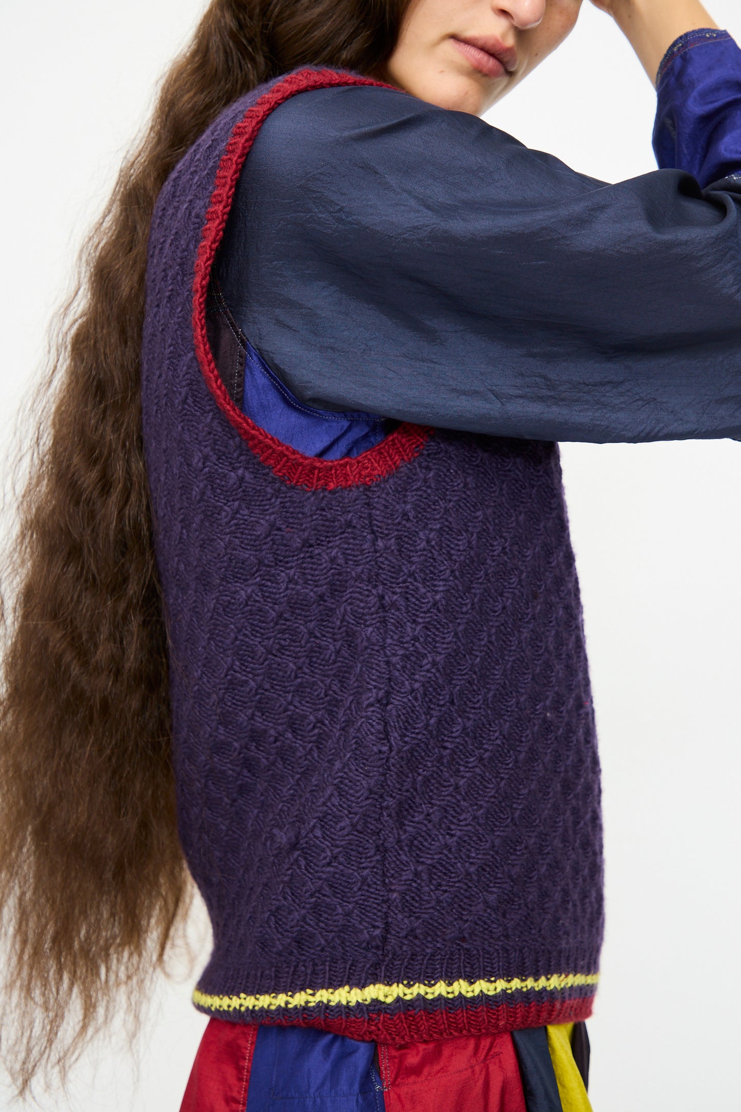 A person is shown from the side wearing the Injiri Merino Wool Sweater Vest in Purple, Red and Yellow over a dark blue long-sleeve shirt and a multicolored skirt.