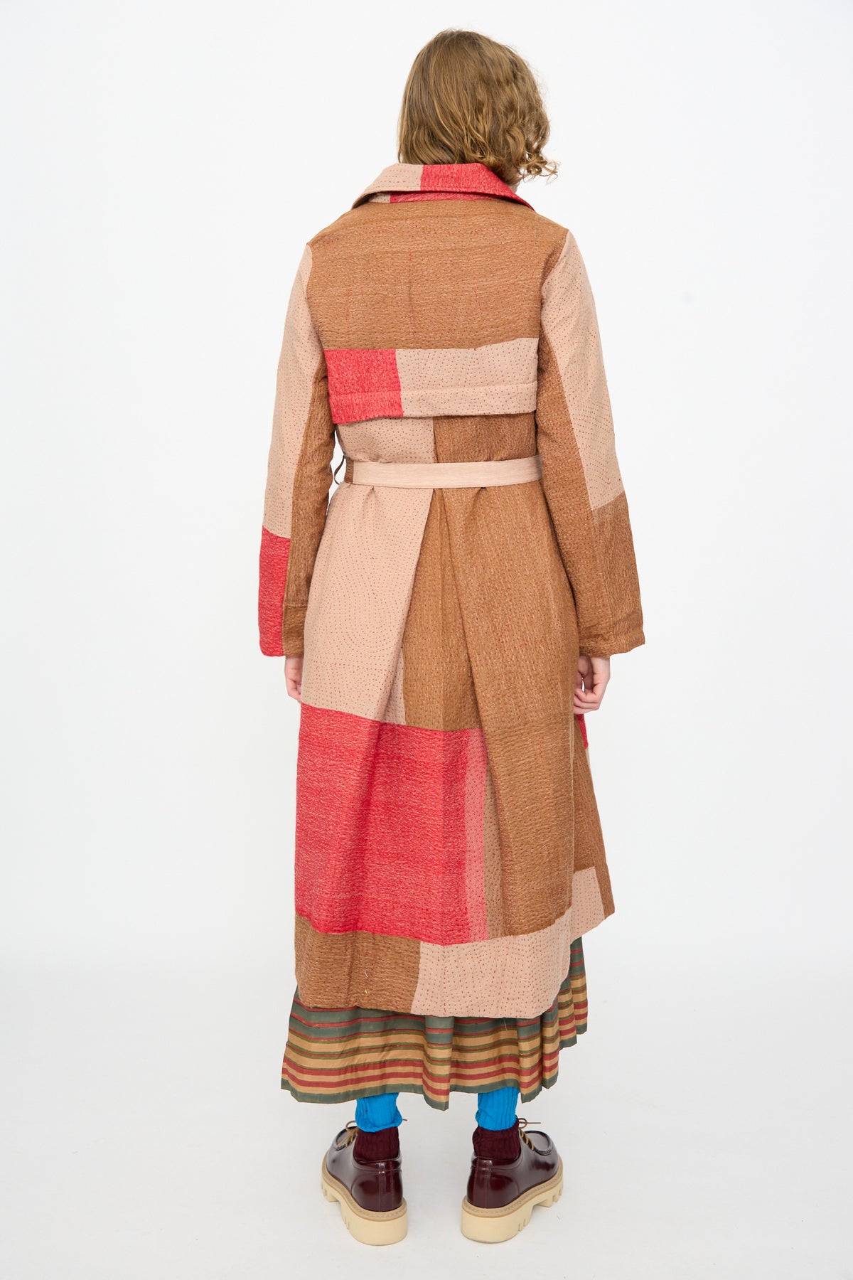 A person faces away from the camera, wearing the Injiri Qasab Wool and Silk Jacket in Red and Brown Multi—a luxurious patchwork coat crafted from a wool-silk blend—paired with a striped skirt, blue socks, and platform shoes, standing against a plain white background.