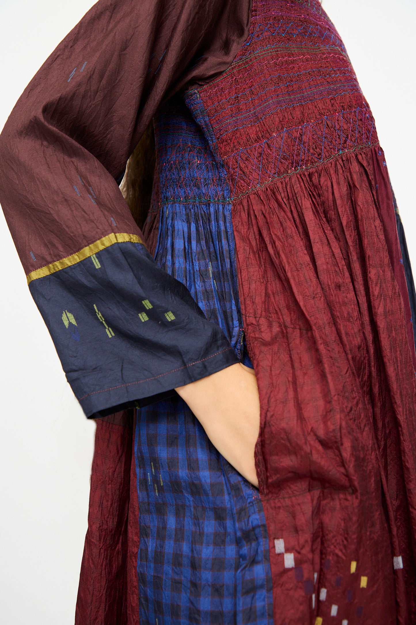 A person wears the Injiri Silk Dress in Red and Blue Multi, featuring long sleeves, an embroidered bodice, patterned maroon and navy fabrics, side view with one hand in a pocket.