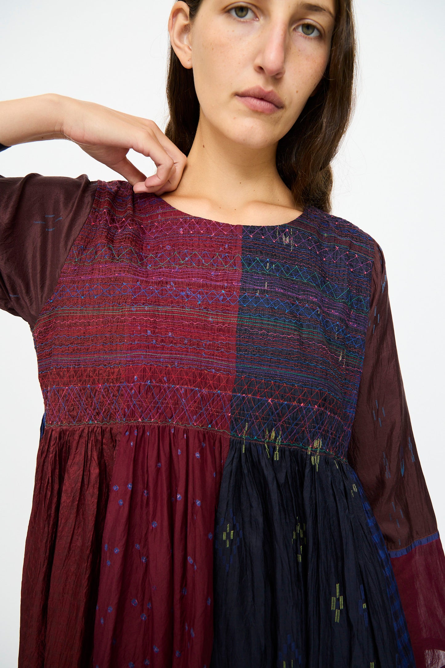 A woman with long brown hair and a neutral expression wears the Injiri Silk Dress in Red and Blue Multi, featuring loose long sleeves, red and dark blue patchwork, and an intricately embroidered bodice.