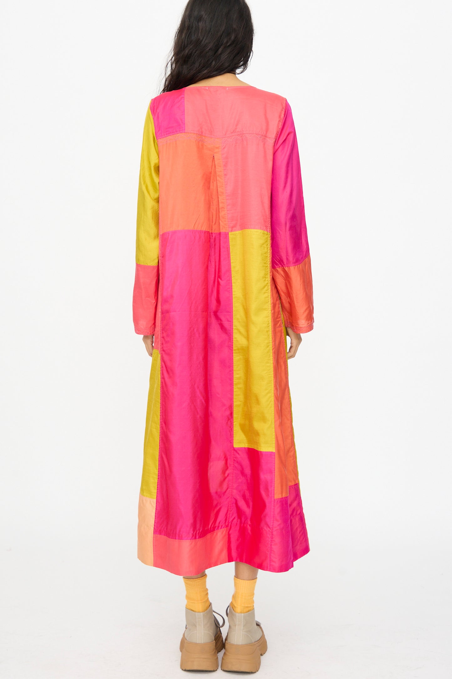 A person faces away wearing the Injiri Silk Dyed Dress in Yellow Pink Multi—a vibrant patchwork design with long sleeves—paired with tan platform shoes and yellow socks.