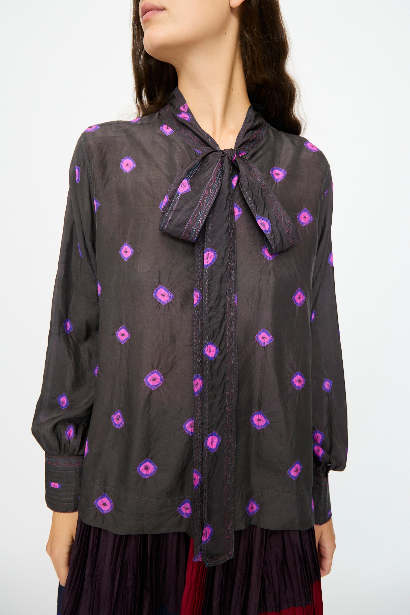 A woman wears the Injiri Silk Shirt in Purple, featuring sheer silk fabric, long sleeves, geometric patterns, and a tied neck bow.