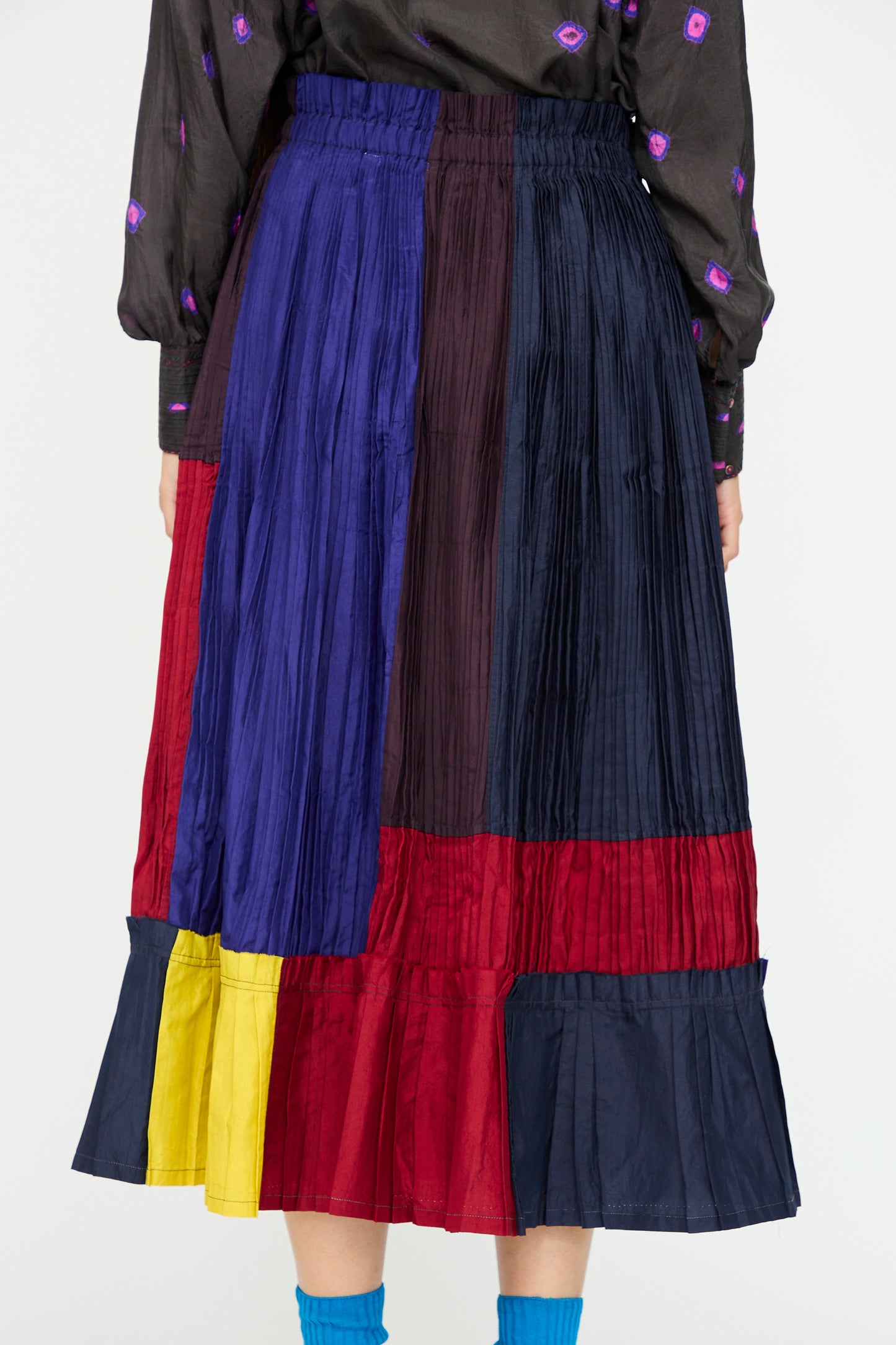 A person wearing the Injiri Taffeta Silk Skirt in Multi—a vibrant, pleated color-block midi—paired with a dark patterned top and blue socks.