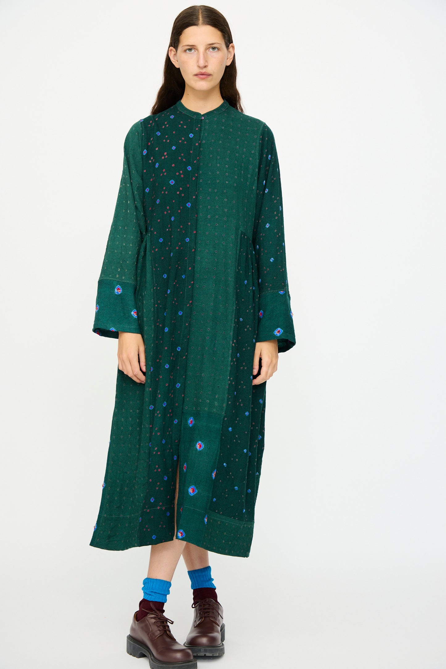 A model wears the Injiri Wool Dress in Green, featuring blue and purple patchwork accents, with blue socks and brown shoes, standing against a plain white background.
