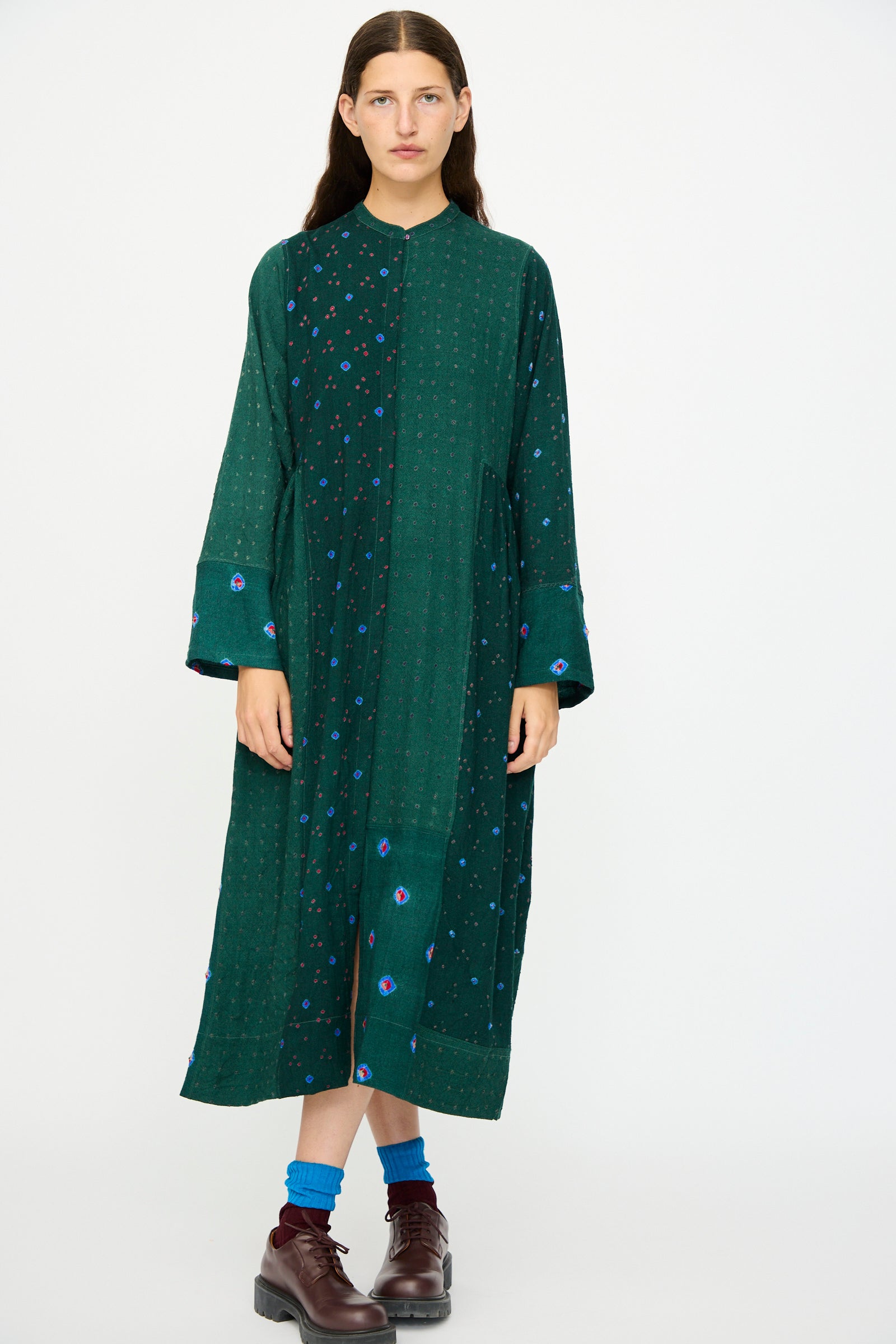 A model wears the Injiri Wool Dress in Green, featuring blue and purple patchwork accents, with blue socks and brown shoes, standing against a plain white background.