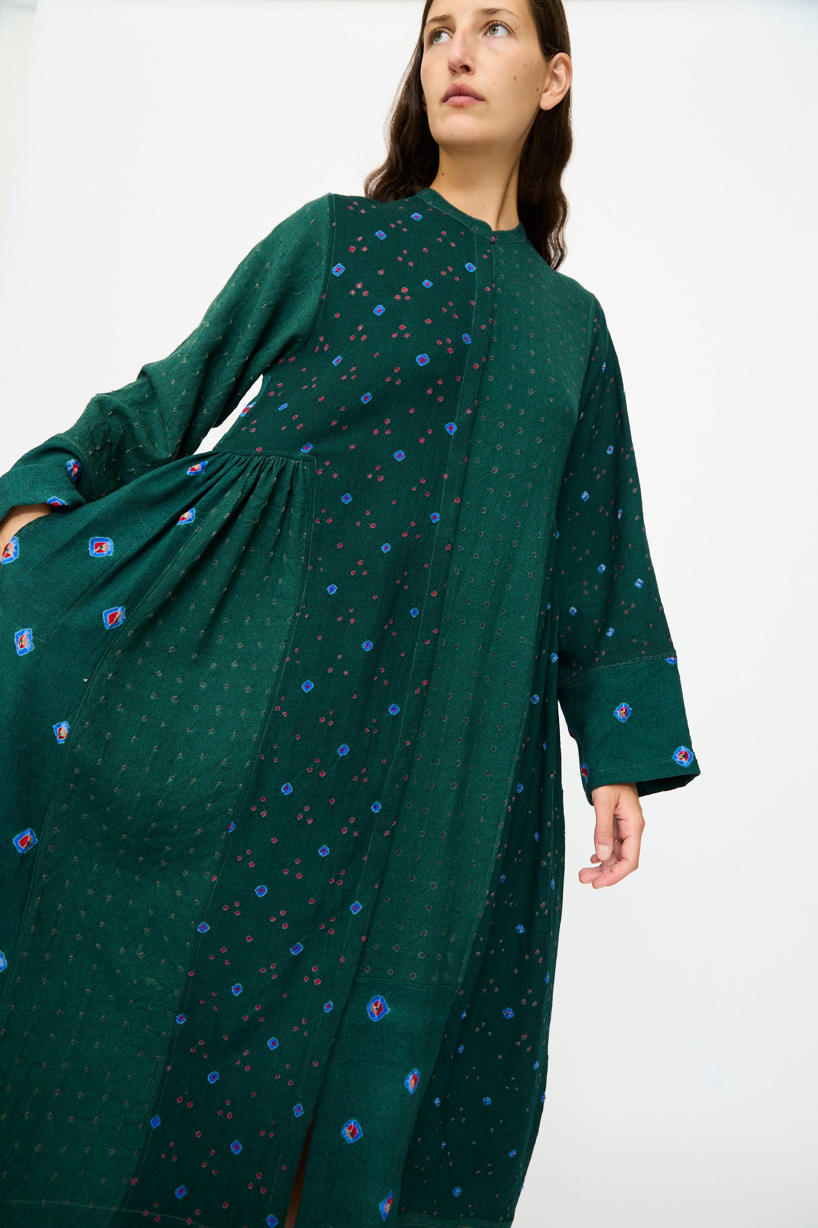 A woman stands against a plain white background wearing the Injiri Wool Dress in Green, featuring long sleeves and subtle blue and red patchwork patterns.