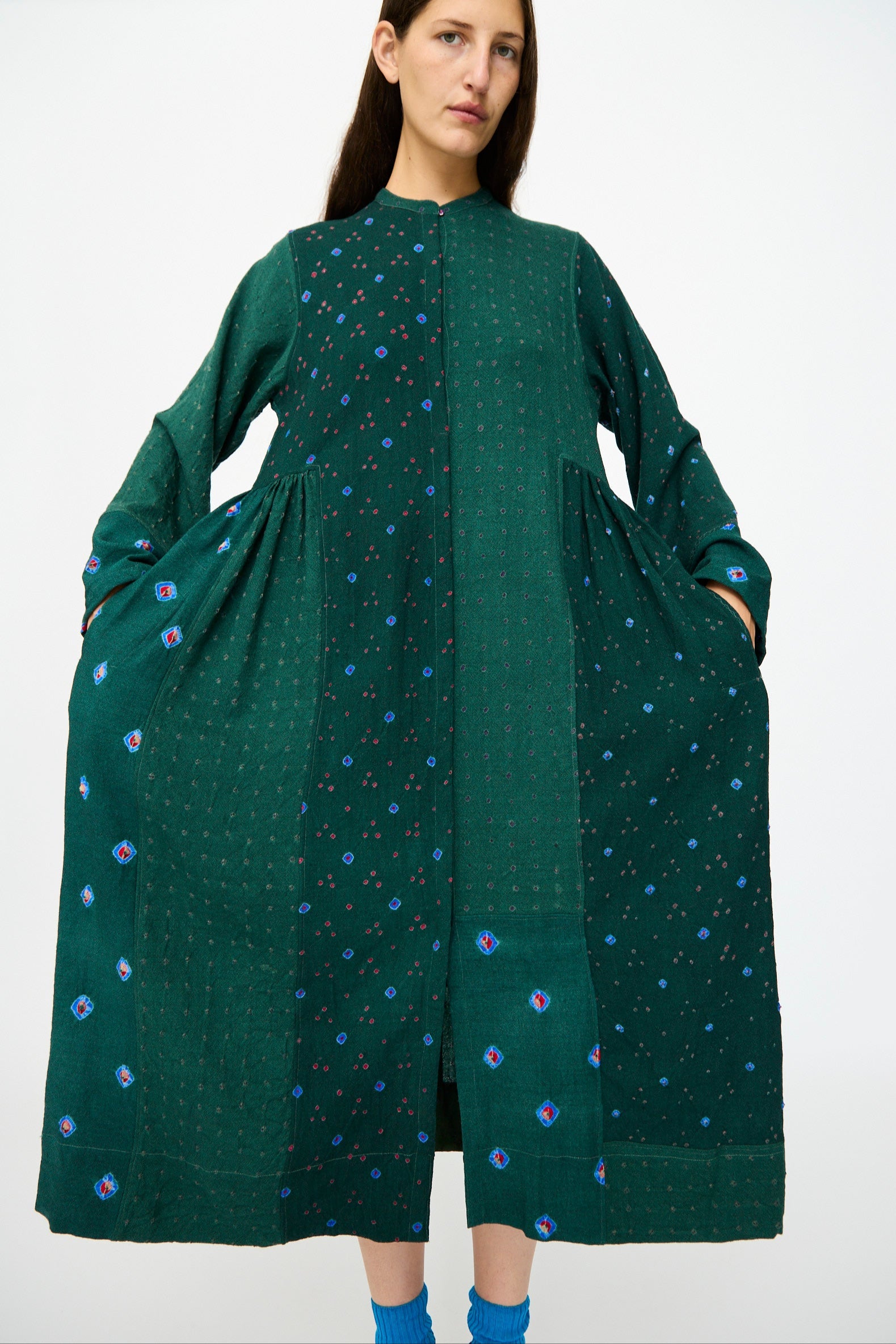 A woman faces forward against a plain white background wearing the Injiri Wool Dress in Green, a long patchwork style featuring pockets, paired with blue socks.
