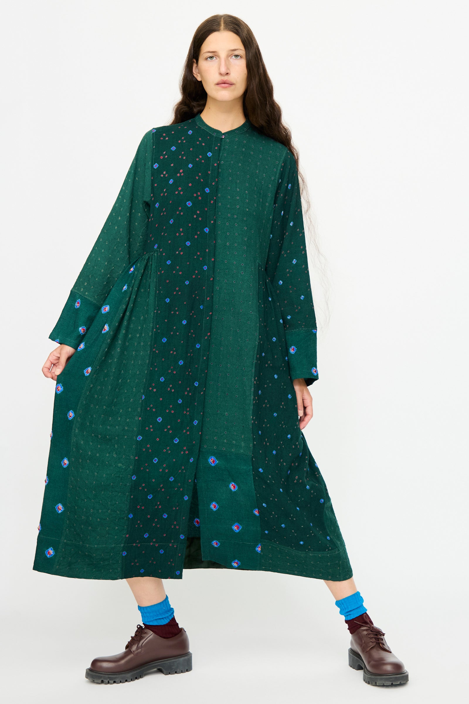 A woman wears the Injiri Wool Dress in Green, featuring long sleeves and a floral pattern, paired with brown shoes and blue socks, standing against a plain white background.