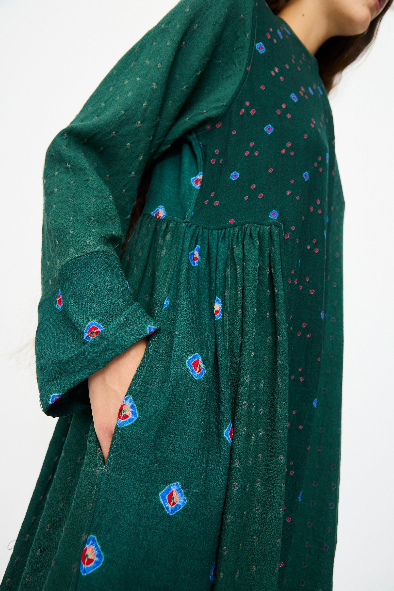 Dressed in Injiri’s Wool Dress in Green, featuring long sleeves and red and blue floral embroidery, this person stands with one hand tucked in the pocket.