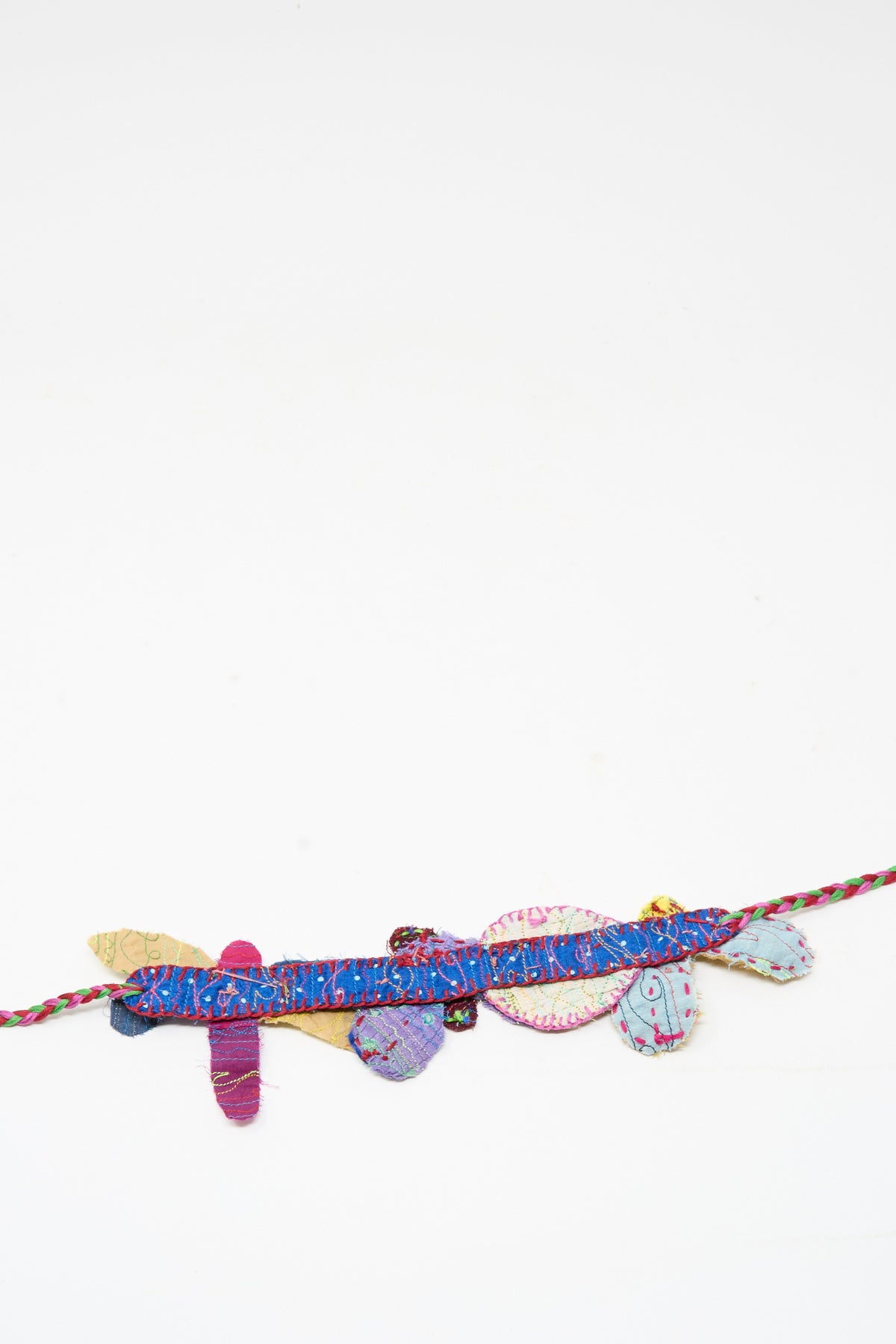 The Wool Silk Neckband in Multi by Injiri is a thin, multicolored braided string adorned with small fabric shapes, evoking the look of a delicate flower patch neckband, and is displayed against a plain white background.
