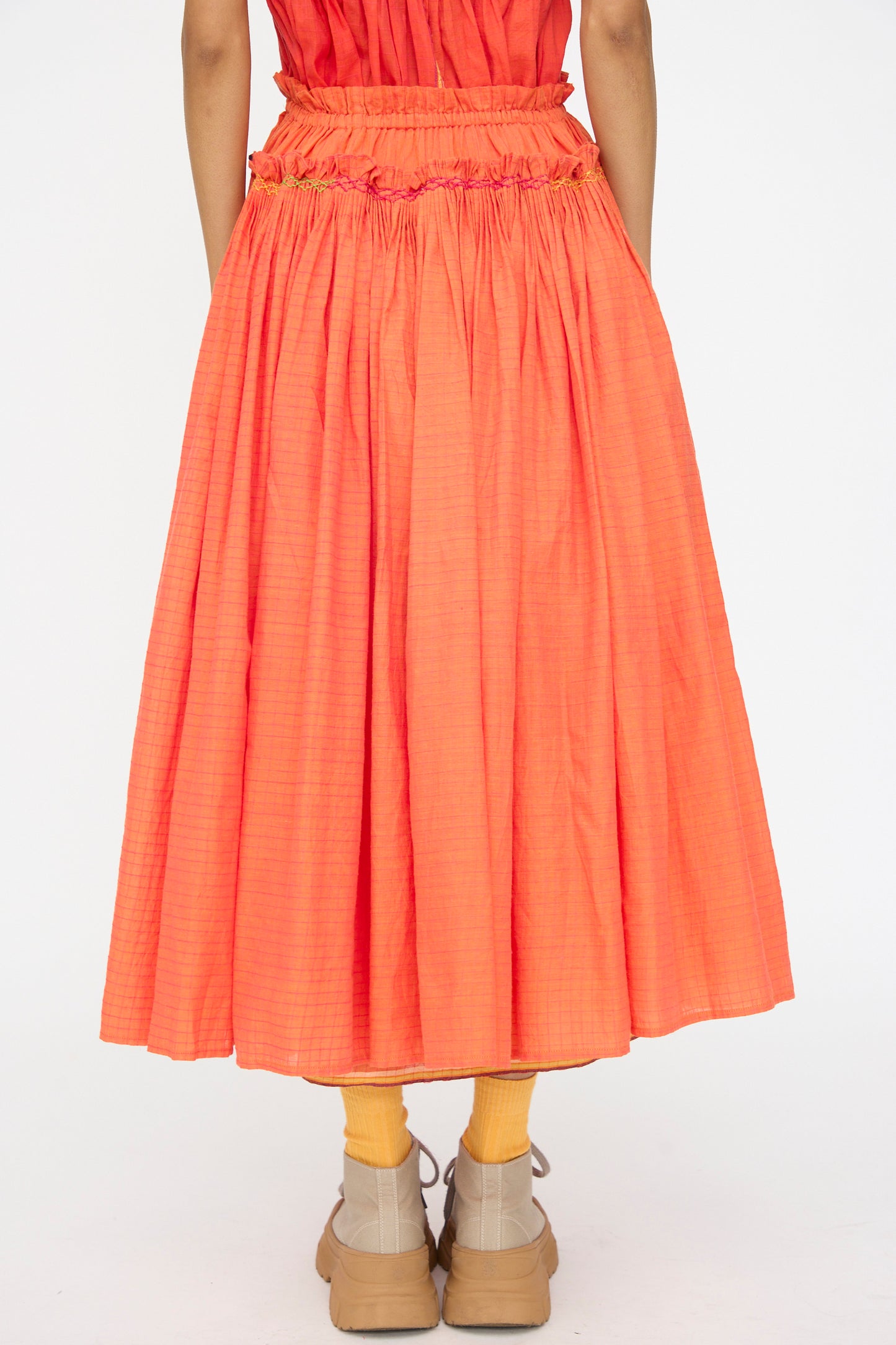 Model shown from the back wearing Injiri’s Woven Cotton Skirt in Orange, featuring pleats and a gathered waistband, paired with yellow socks and tan shoes against a plain white background.
