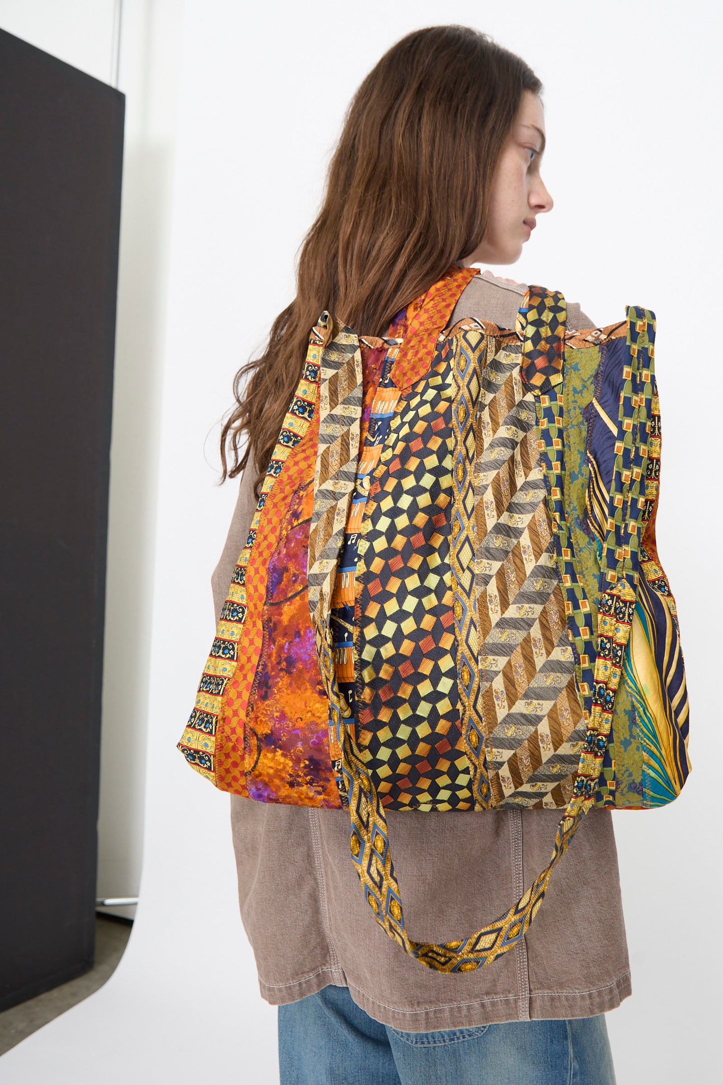 A person with long brown hair carries the Intensity Ties Bag in Orange Multi over their shoulder, paired with a brown shirt and blue jeans.