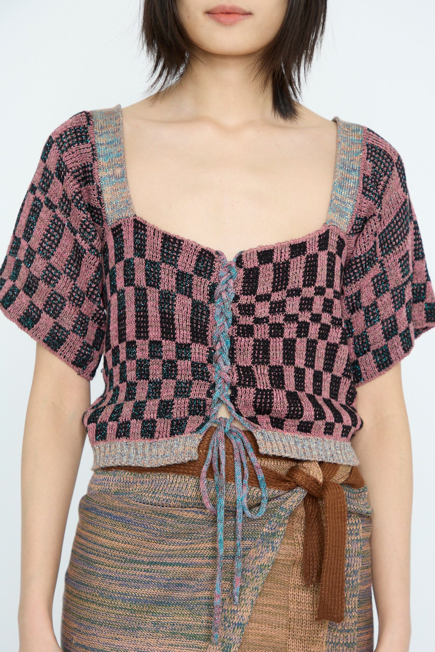 A person models the Intensity Wing Corset in Pink, featuring a pink and black checkered pattern with metallic yarn details, paired with a multicolored wrap skirt against a plain background.