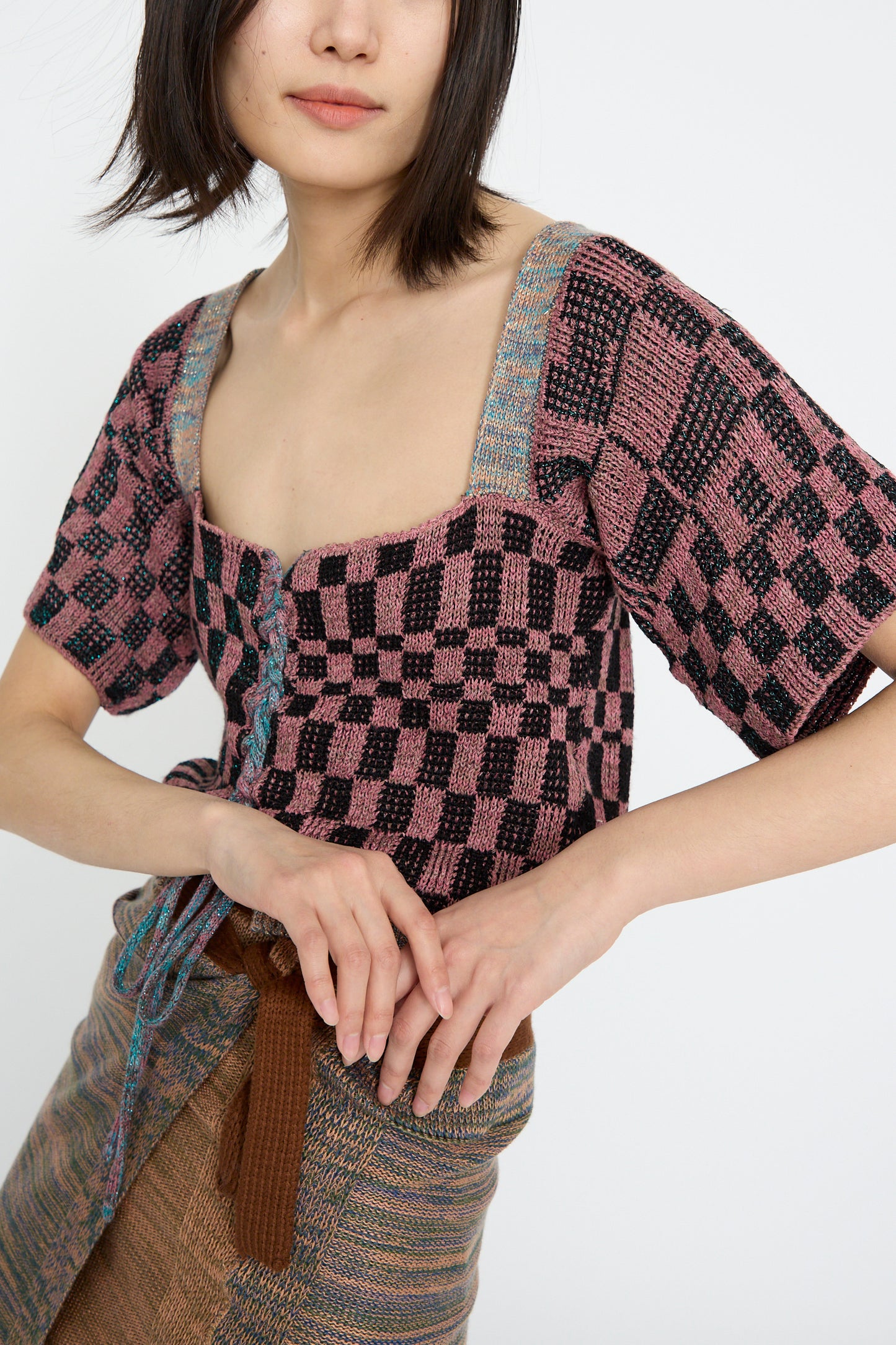 A woman stands against a plain background, wearing the Intensity Wing Corset in Pink and a woven skirt, arms in front.