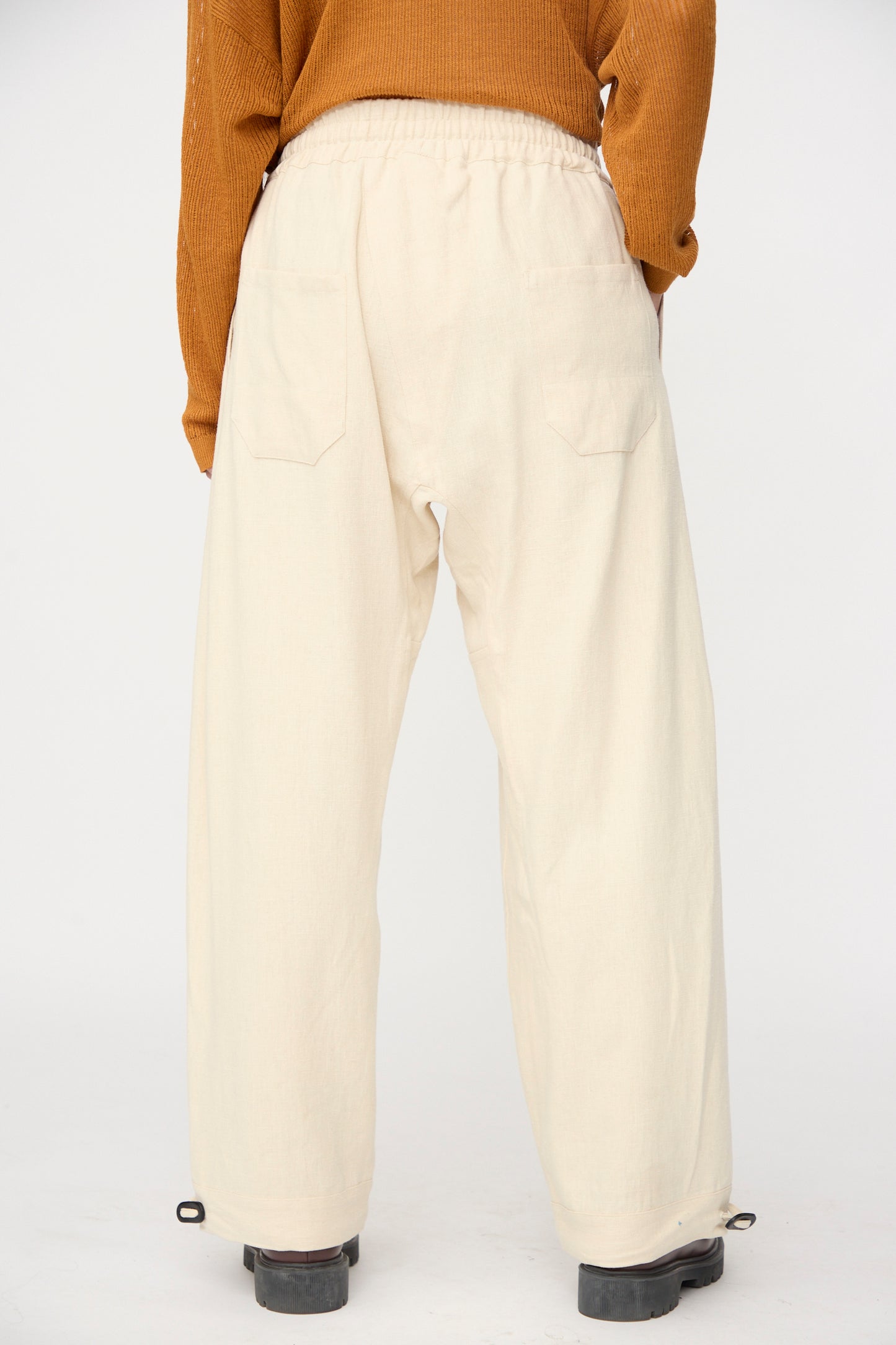 A person stands with their back to the camera in Jan-Jan Van Essche Hemp Soft Canvas Trousers #91 in Kinari, featuring a loose fit and elasticated waistband, paired with a brown sweater against a plain background.