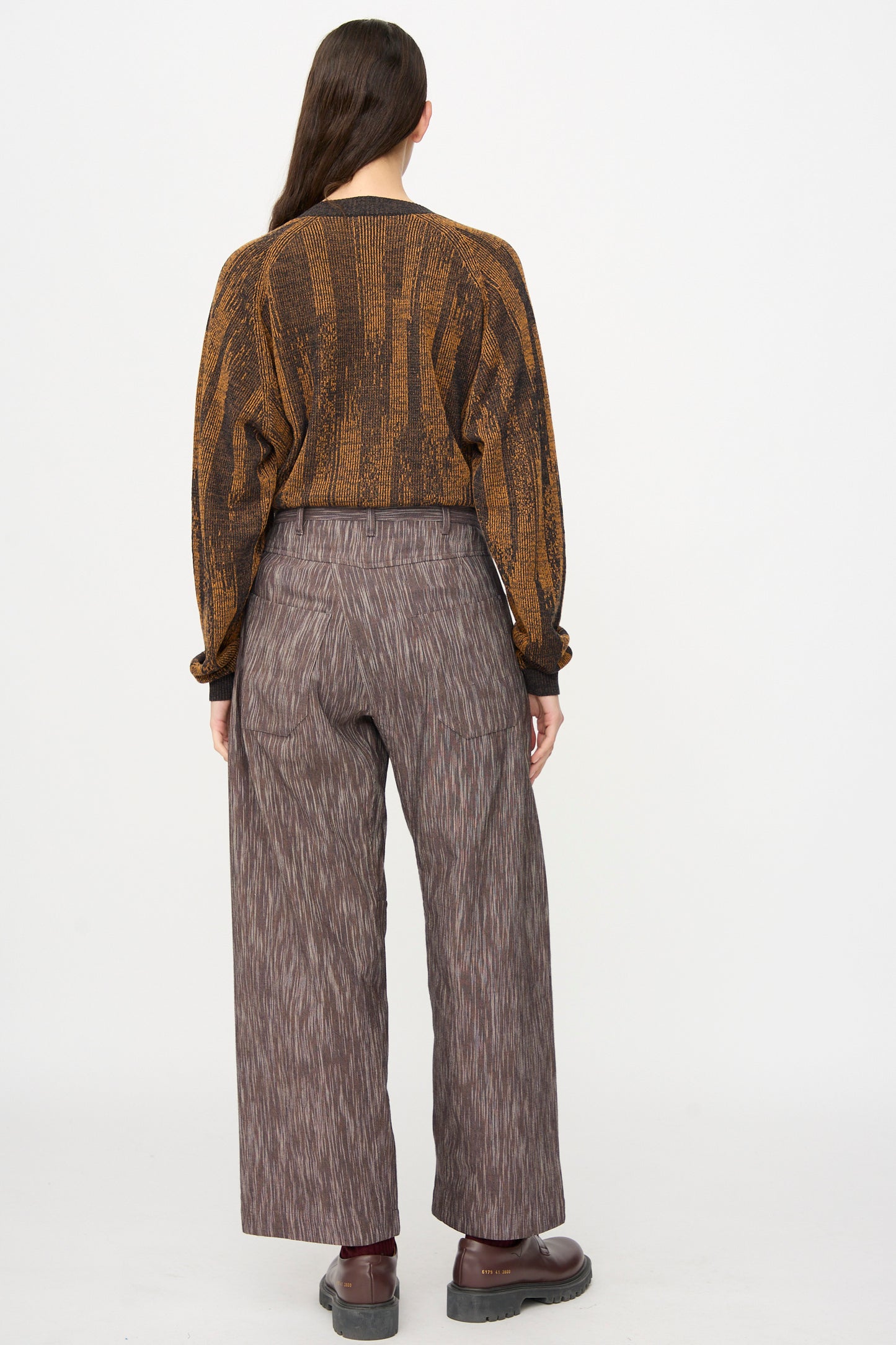 A person stands facing away, wearing a brown patterned sweater, Jan-Jan Van Essche Kasuri Cotton Silk Trousers #90 in Old Lavender, and dark shoes against a plain white background.
