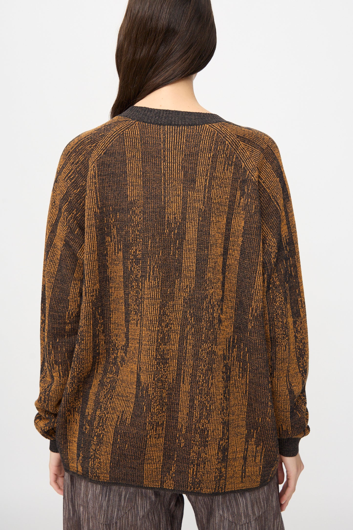 Person stands with their back to the camera, wearing Jan-Jan Van Essche's Linen Cotton Kasuri Knit #77 in Black Brown Orange, paired with brown patterned pants against a plain background.