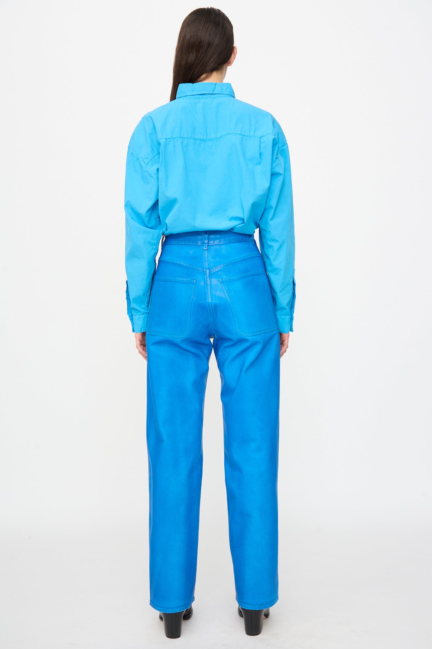 A person stands facing away, wearing Jesse Kamm's 225 Jeans in Electric Blue Lacquer, paired with a long-sleeve blue shirt and black heeled shoes against a plain white background.