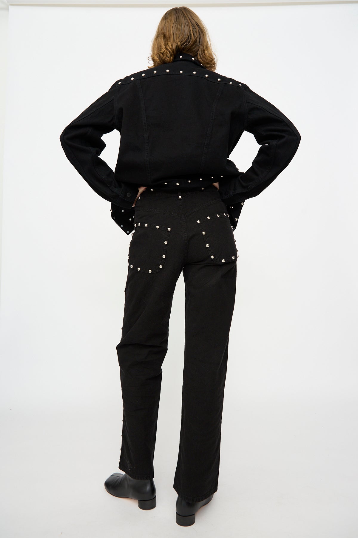 A person stands with hands on hips, wearing Jesse Kamm’s 225′s in Black with Studs—high-rise pants crafted from organic cotton canvas and embellished with metal studs—paired with a black denim jacket, facing away from the camera against a plain white background.