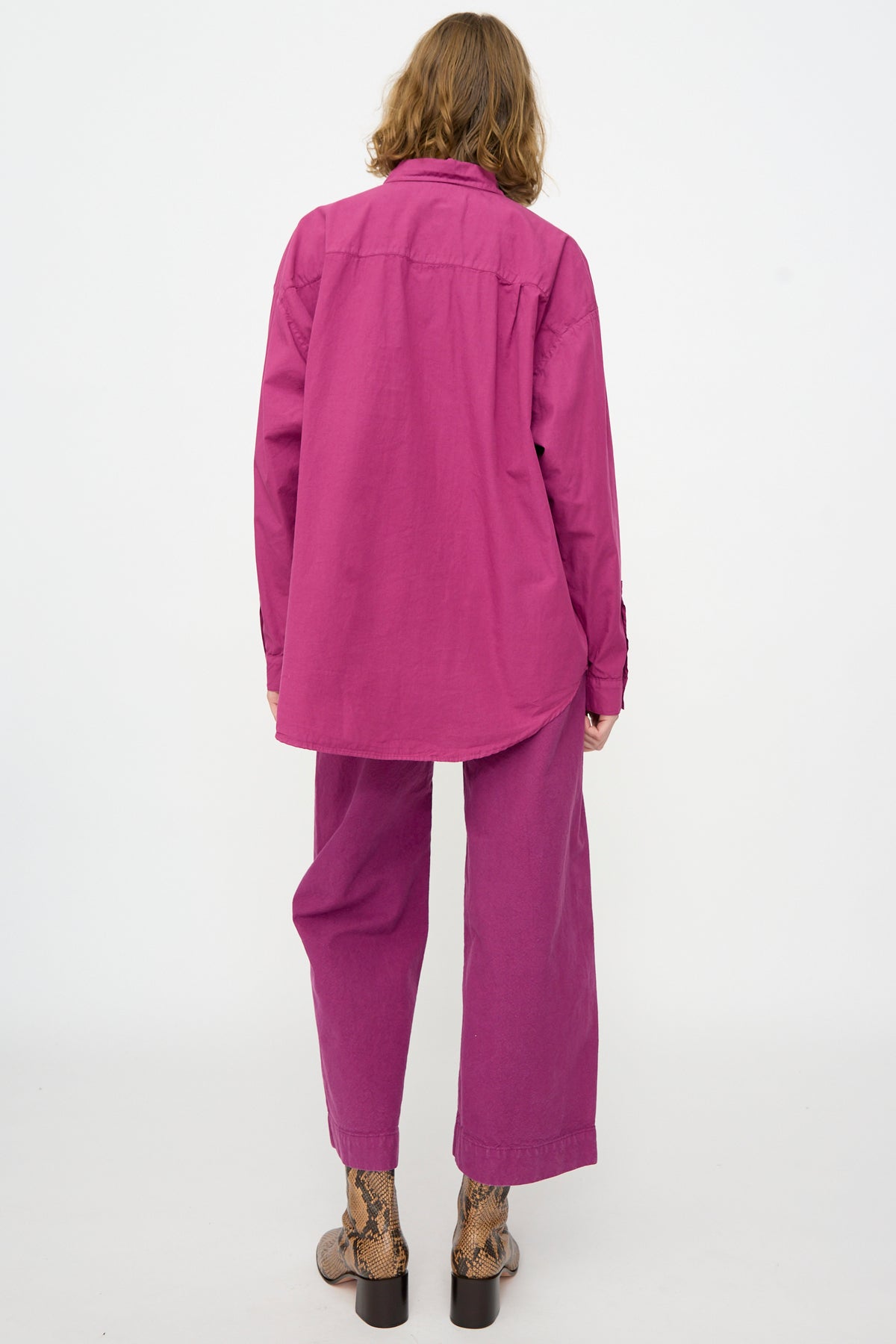 A person stands facing away from the camera against a plain white background, wearing the Jesse Kamm Alberta Shirt in Beet in an oversized fit, paired with matching pants and snake-patterned shoes.