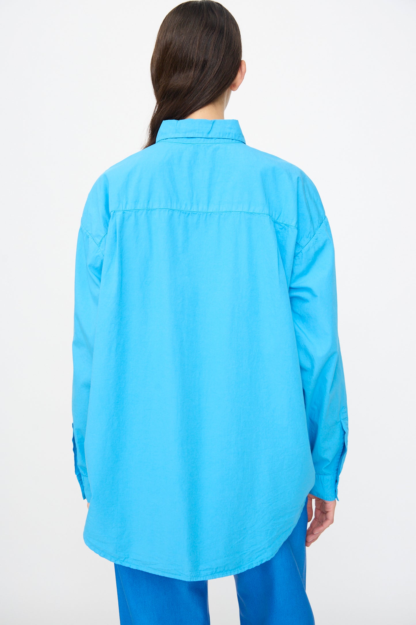 Model with long dark hair, seen from the back against a white background, wears the Jesse Kamm Alberta Shirt in Pool Blue—an oversized organic cotton design—paired with blue pants.