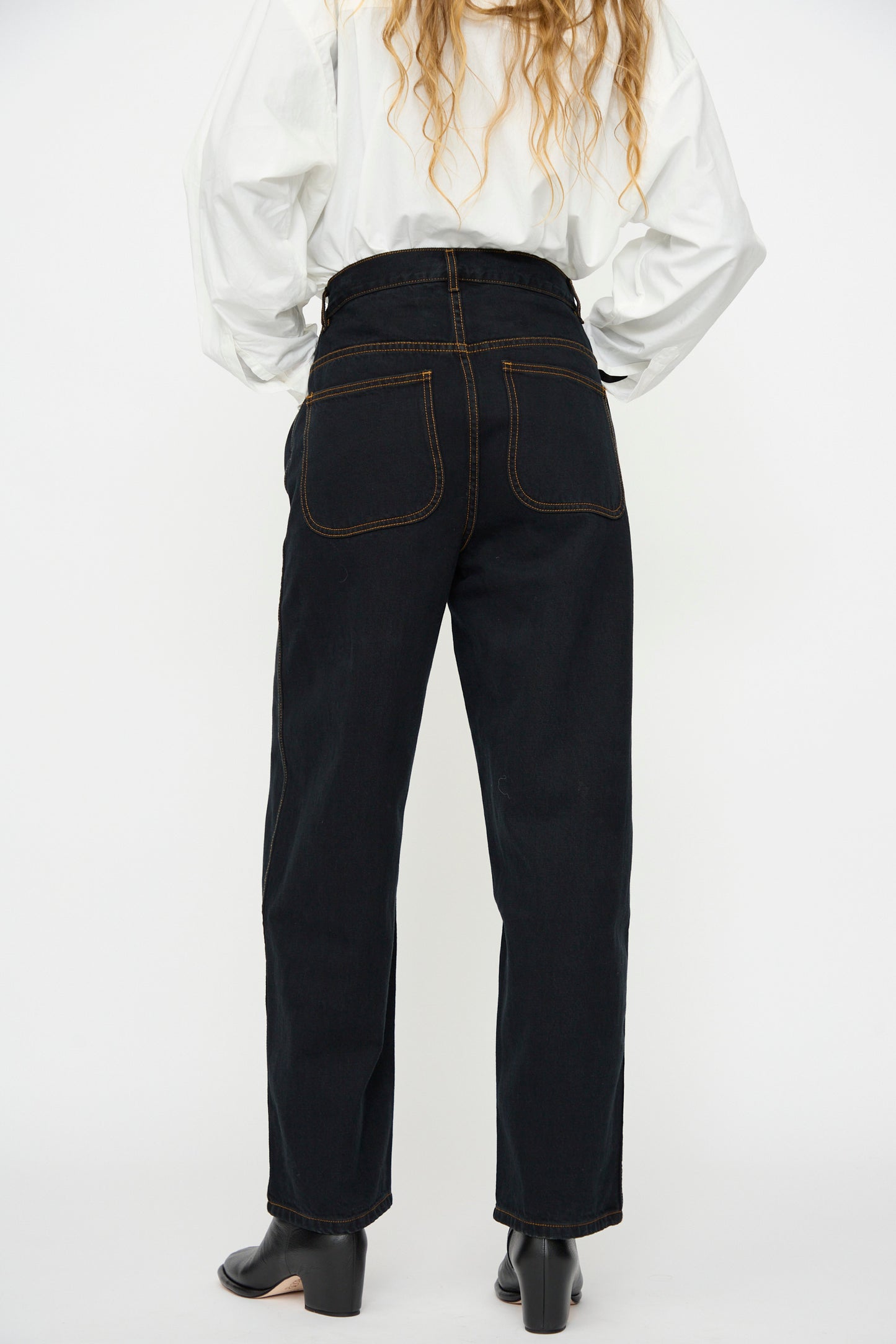 A person shown from the back wears a white long-sleeve shirt, Jesse Kamm’s California Wide in Black Overdye jeans with brown stitching, and black heeled shoes.