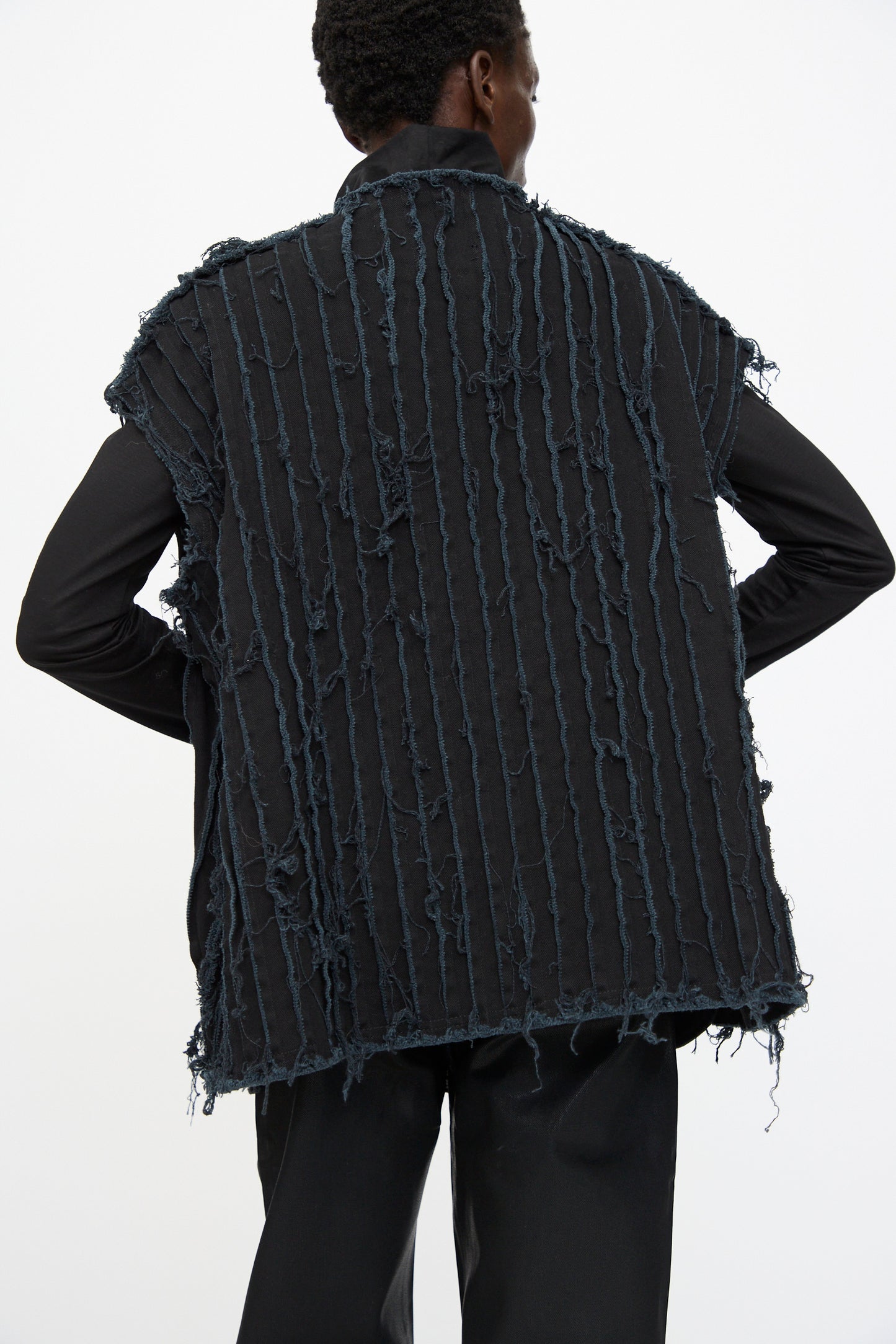 A person is seen from behind wearing the Jesse Kamm Deadstock 10oz. Denim Baleen Vest in Black over a long-sleeve black shirt and black pants, featuring vertical frayed textured lines.