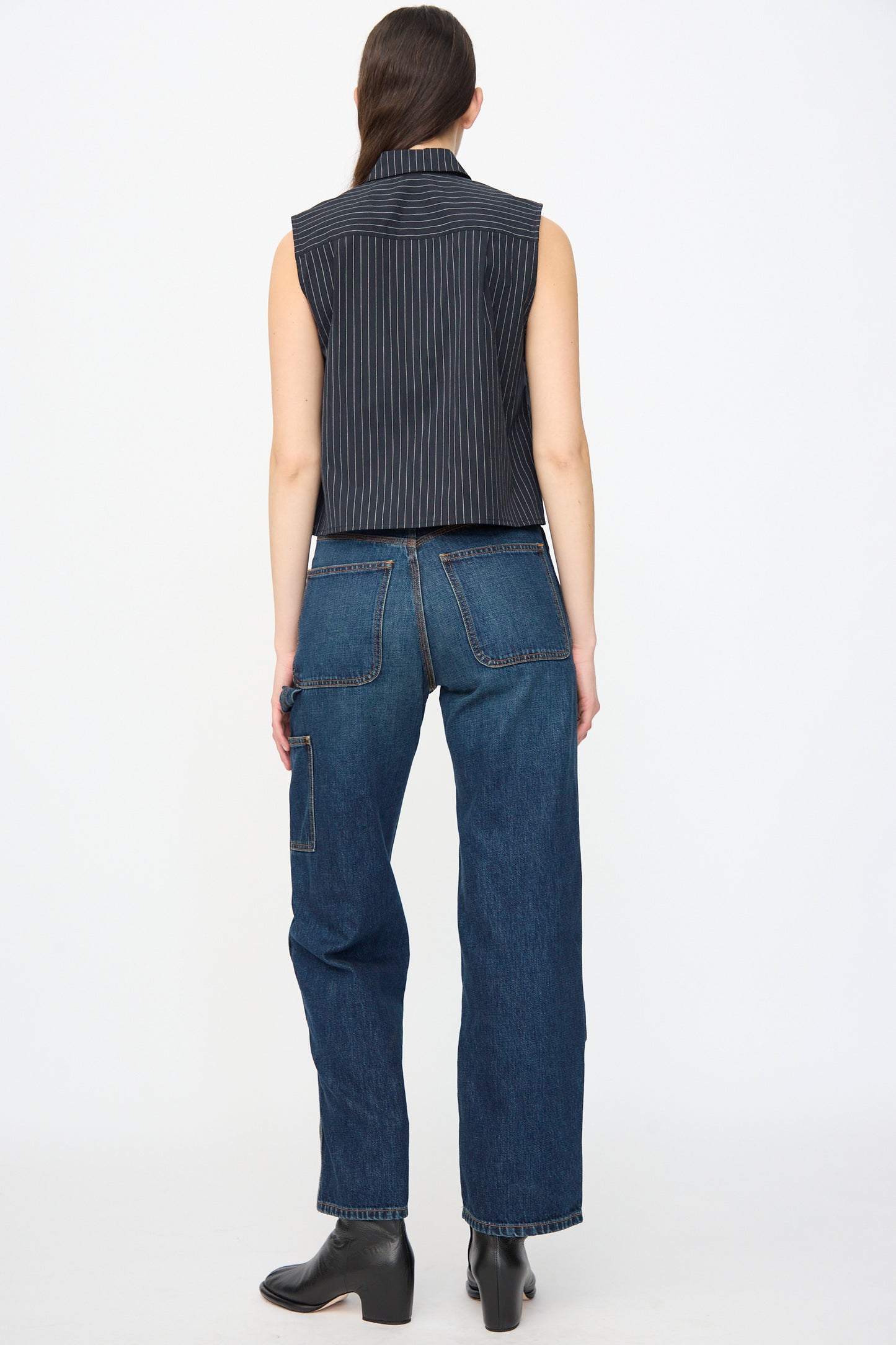 A person stands facing away, wearing a sleeveless pinstriped top, Jesse Kamm Denim Patchwork Handy Pants in Medium Roast with wide legs and large back pockets, paired with black ankle boots.