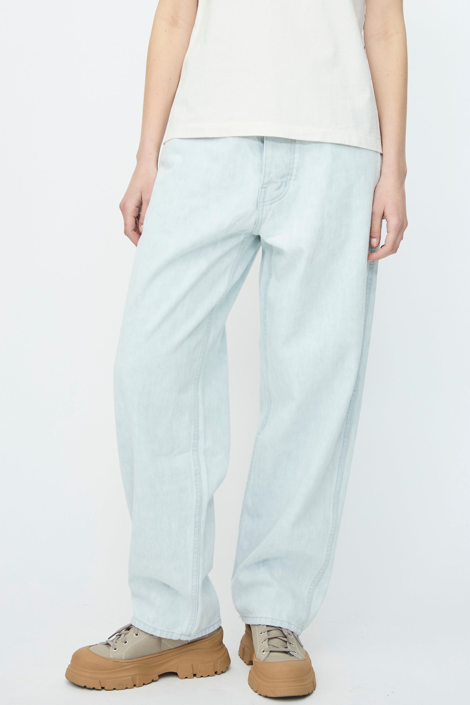 Against a plain backdrop, an individual stands wearing Jesse Kamm's Japanese Denim California Wide in Pale Blue, paired with a white shirt and beige shoes.