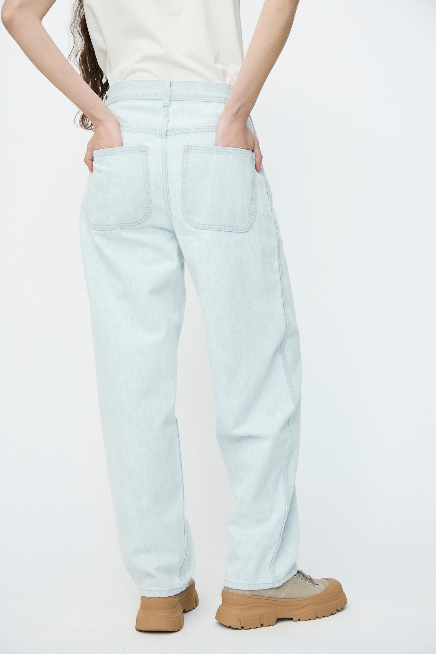 A person in Jesse Kamm's Japanese Denim California Wide in Pale Blue jeans and tan shoes stands with hands in back pockets against a plain background.