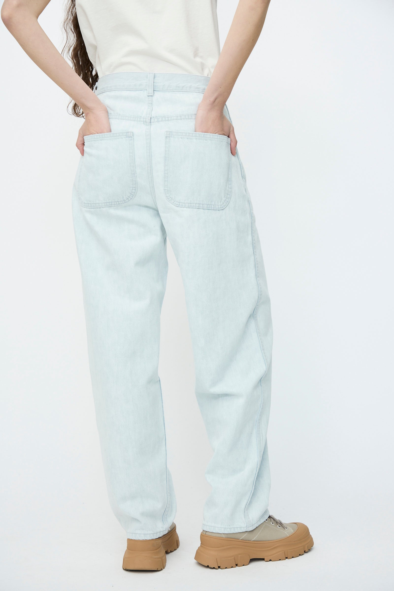 A person in Jesse Kamm's Japanese Denim California Wide in Pale Blue jeans and tan shoes stands with hands in back pockets against a plain background.
