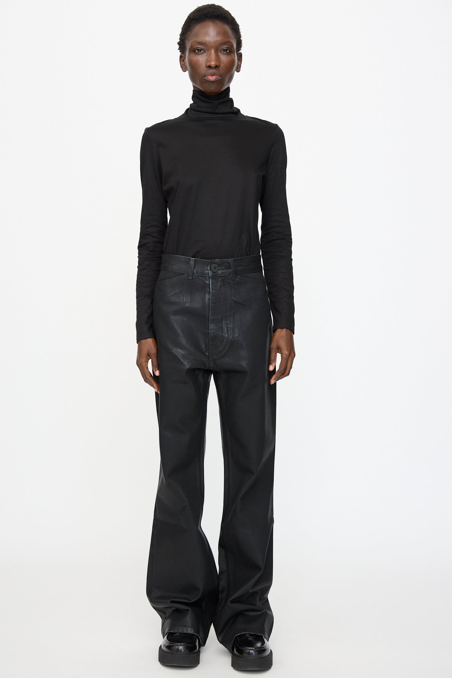 Person standing against a plain background, wearing a black turtleneck top, Jesse Kamm Japanese Denim Les Deux Pants in Black Lacquer, and black shoes.