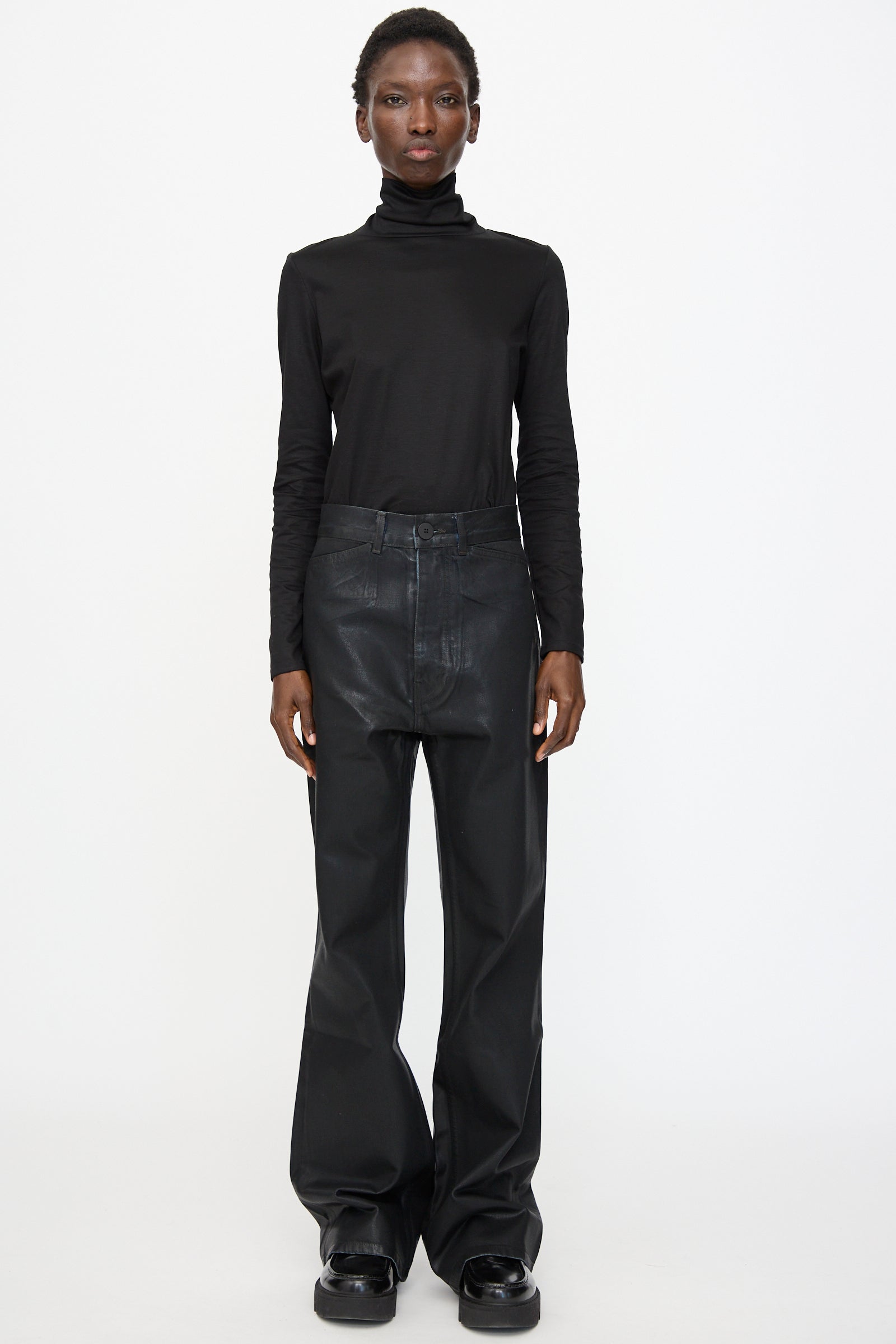 Person standing against a plain background, wearing a black turtleneck top, Jesse Kamm Japanese Denim Les Deux Pants in Black Lacquer, and black shoes.