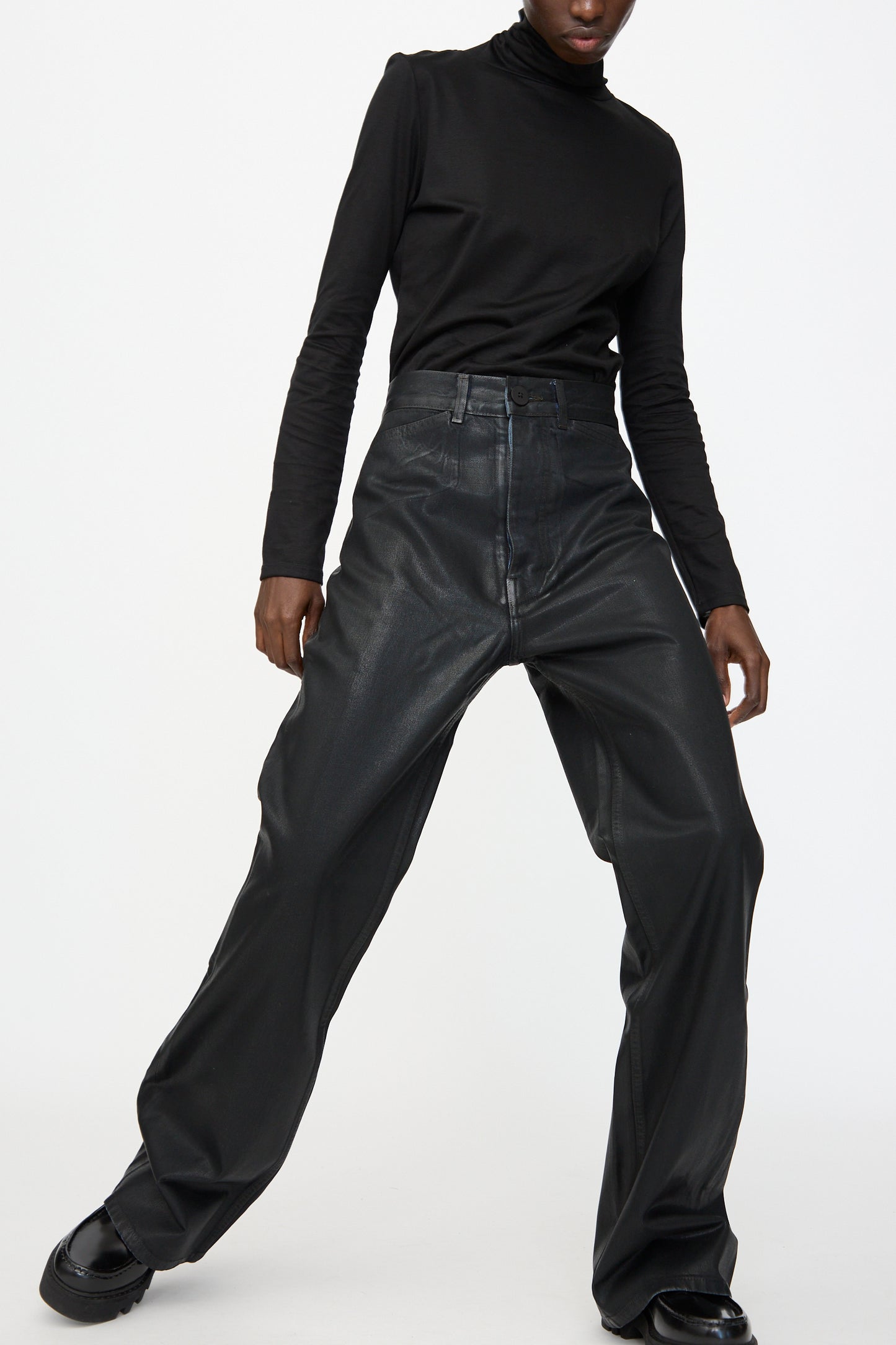 A person wearing a black turtleneck, Jesse Kamm Japanese Denim Les Deux Pants in Black Lacquer, and black shoes stands against a plain white background.