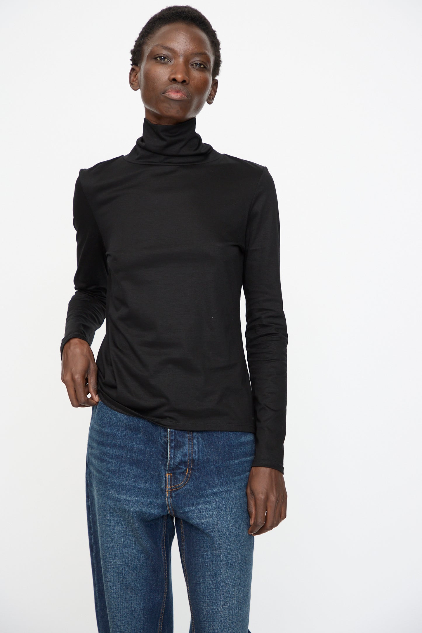 A person stands against a plain white background wearing the Jesse Kamm Japanese Organic Cotton Classic Turtleneck in Black and blue jeans.