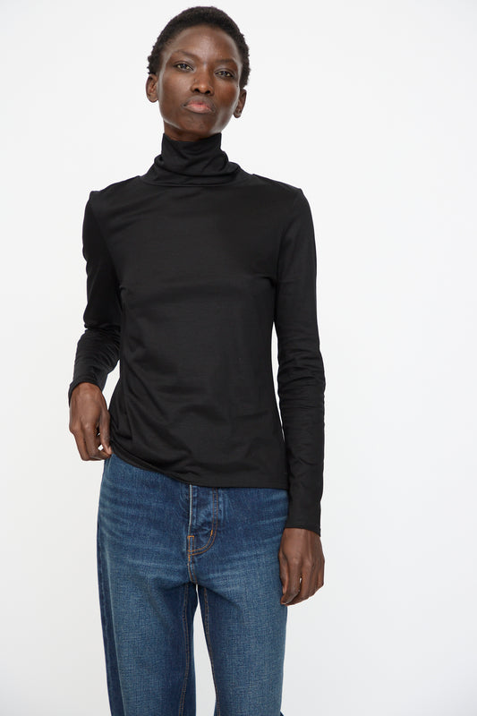 A person stands against a plain white background wearing the Jesse Kamm Japanese Organic Cotton Classic Turtleneck in Black and blue jeans.