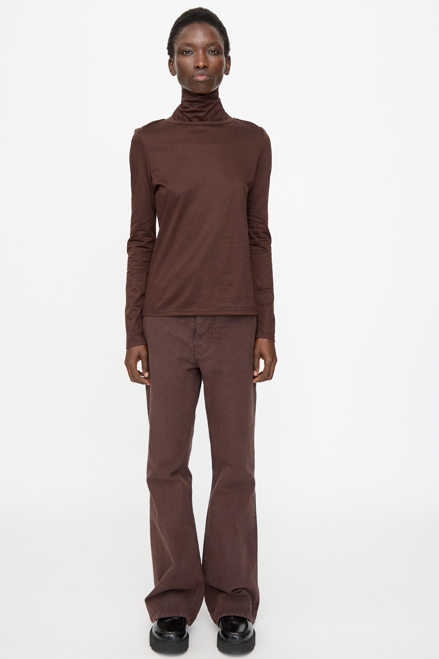 A person stands against a white background wearing the Jesse Kamm Japanese Organic Cotton Classic Turtleneck in Raisin, paired with matching brown pants and black shoes.