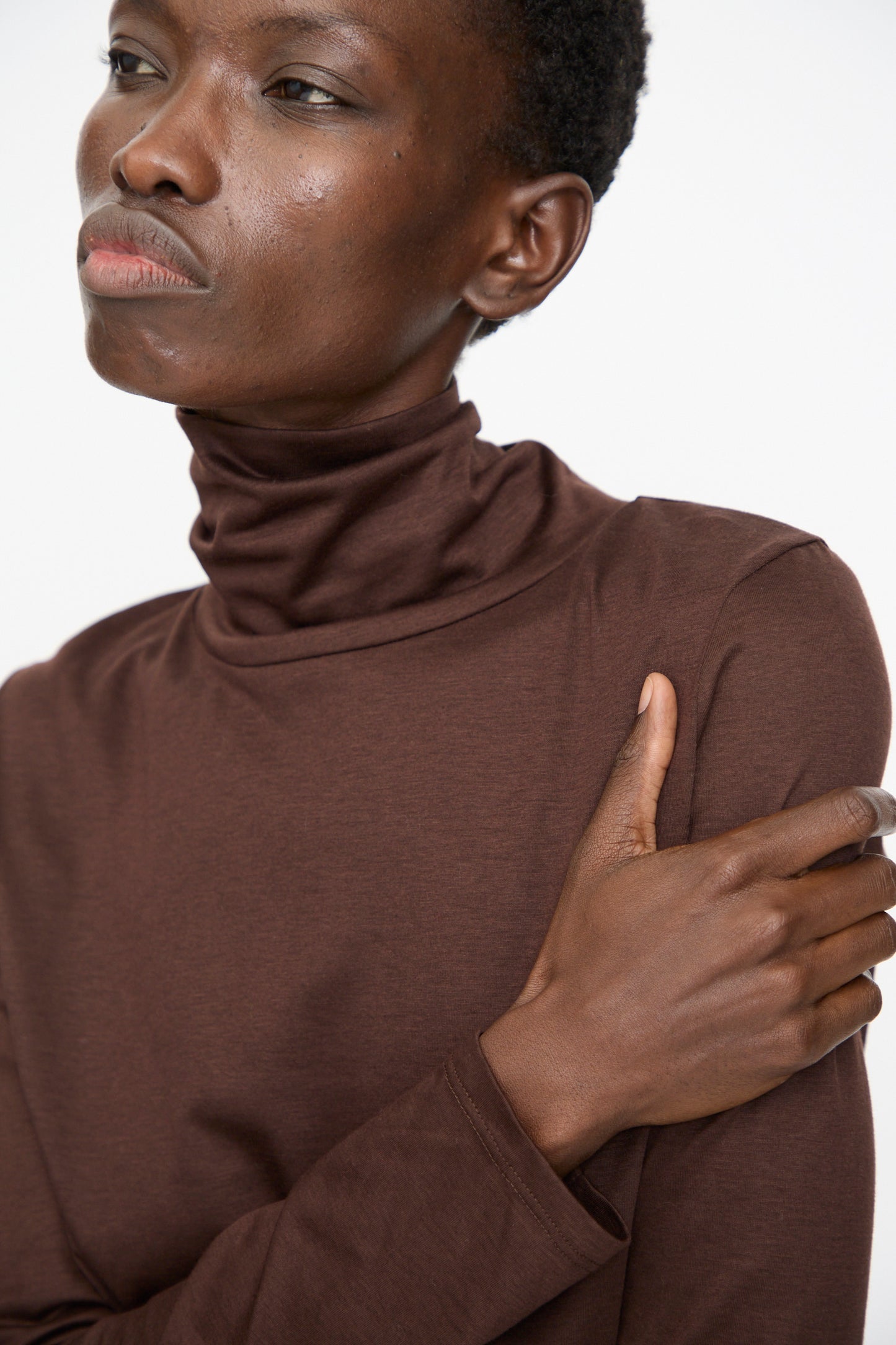 Model wears the Jesse Kamm Japanese Organic Cotton Classic Turtleneck in Raisin, one arm crossed over their chest, gazing slightly to the side against a plain background.