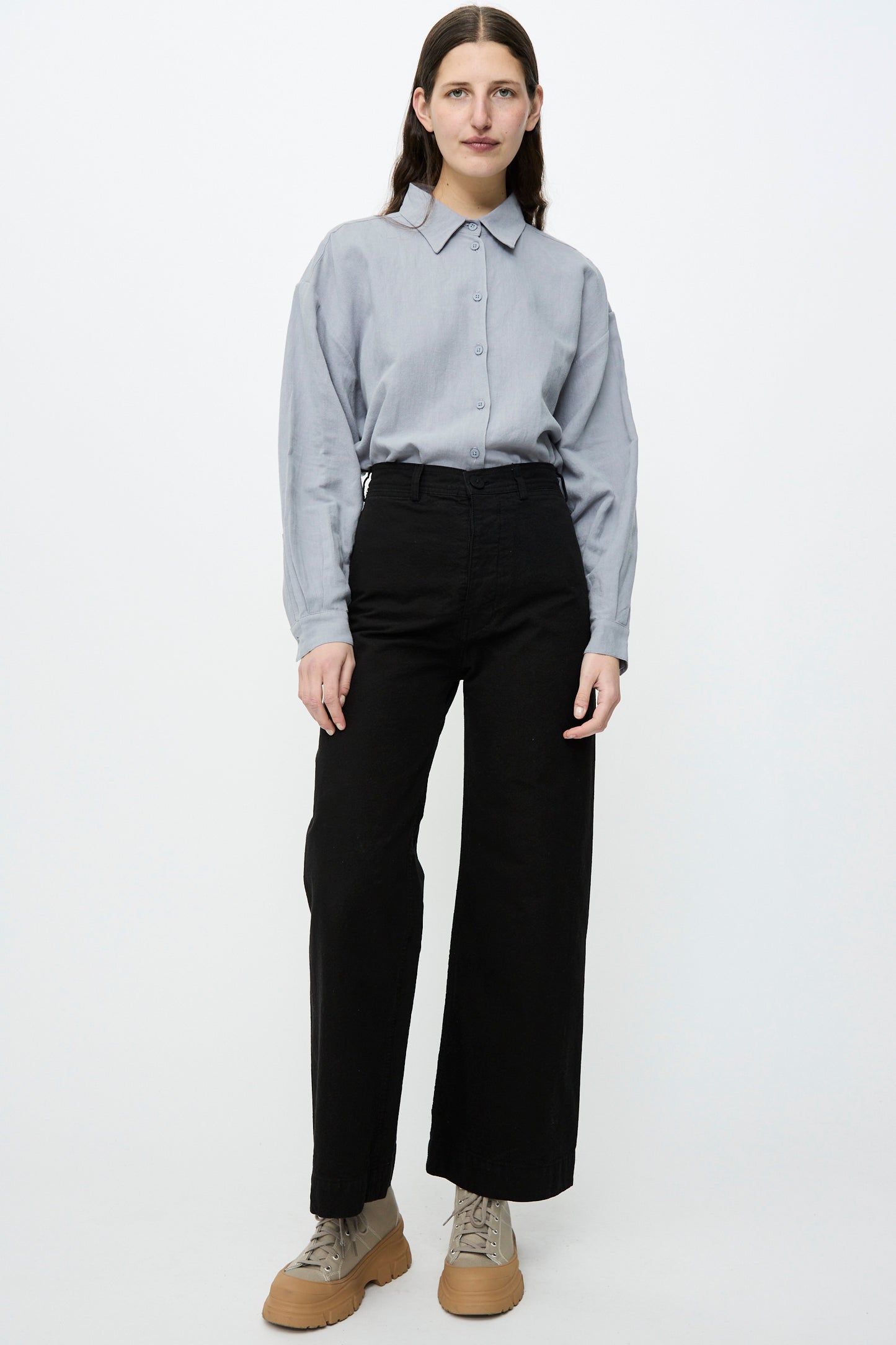 A person stands against a plain white background wearing a light blue button-up shirt, Jesse Kamm's Sailor Pant in Black made of organic cotton canvas, and beige shoes.