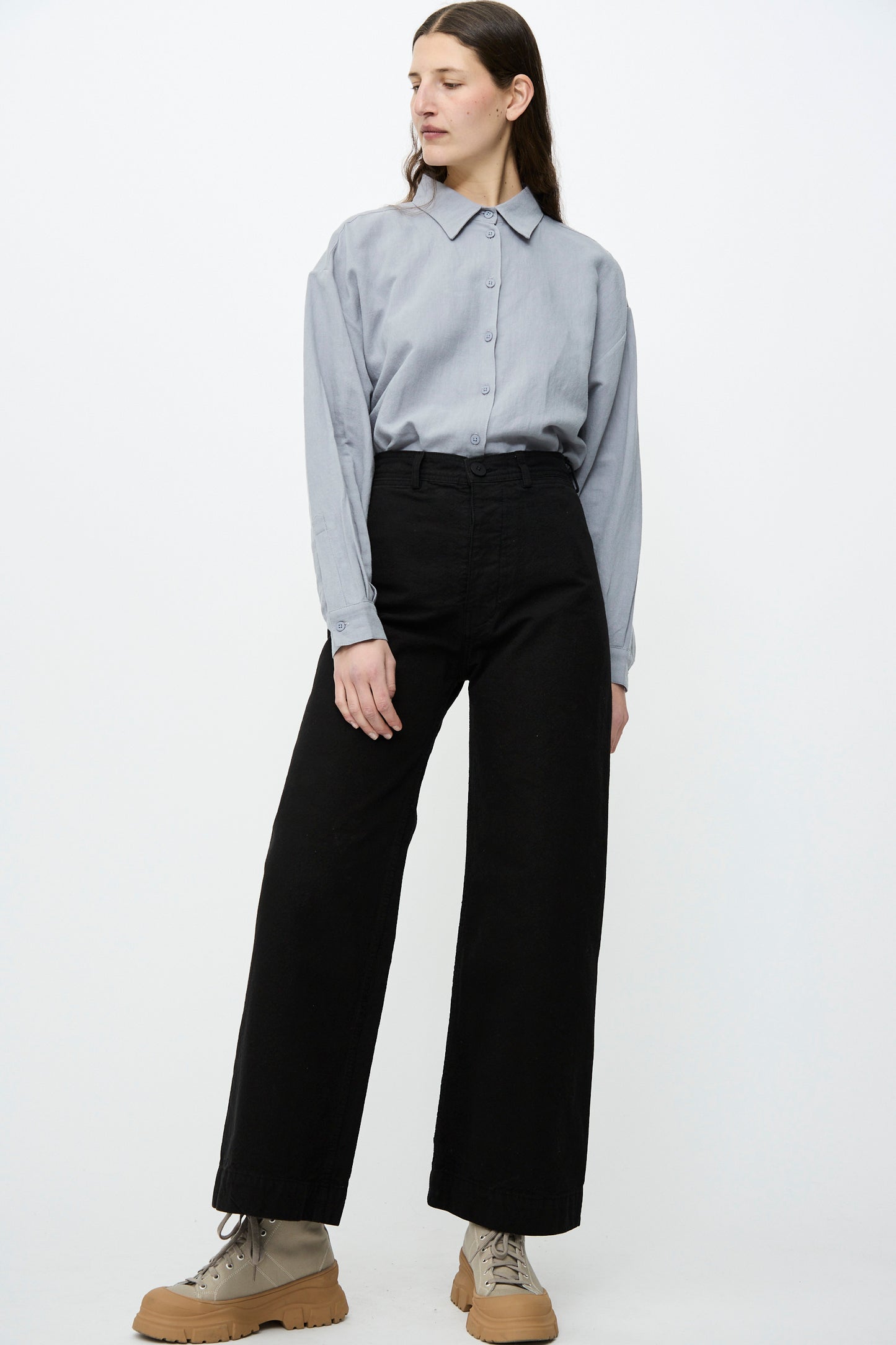 Against a plain backdrop, a person wears Jesse Kamm's Sailor Pant in Black, made from organic cotton canvas. Paired with a light blue button-up and beige boots.