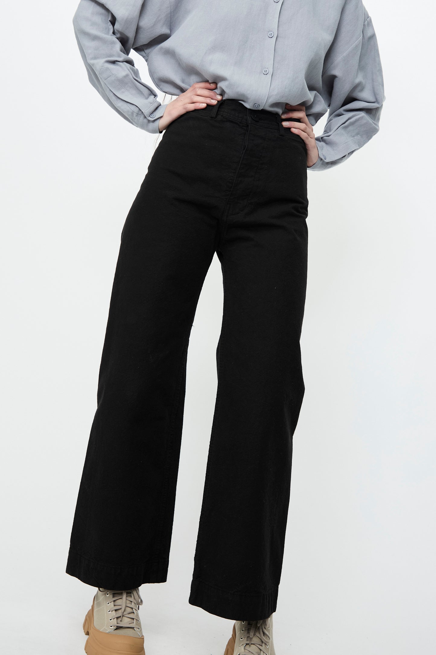 Against a white backdrop, a person poses with hands on hips, wearing a light gray shirt paired with Jesse Kamm's Sailor Pant in Black.
