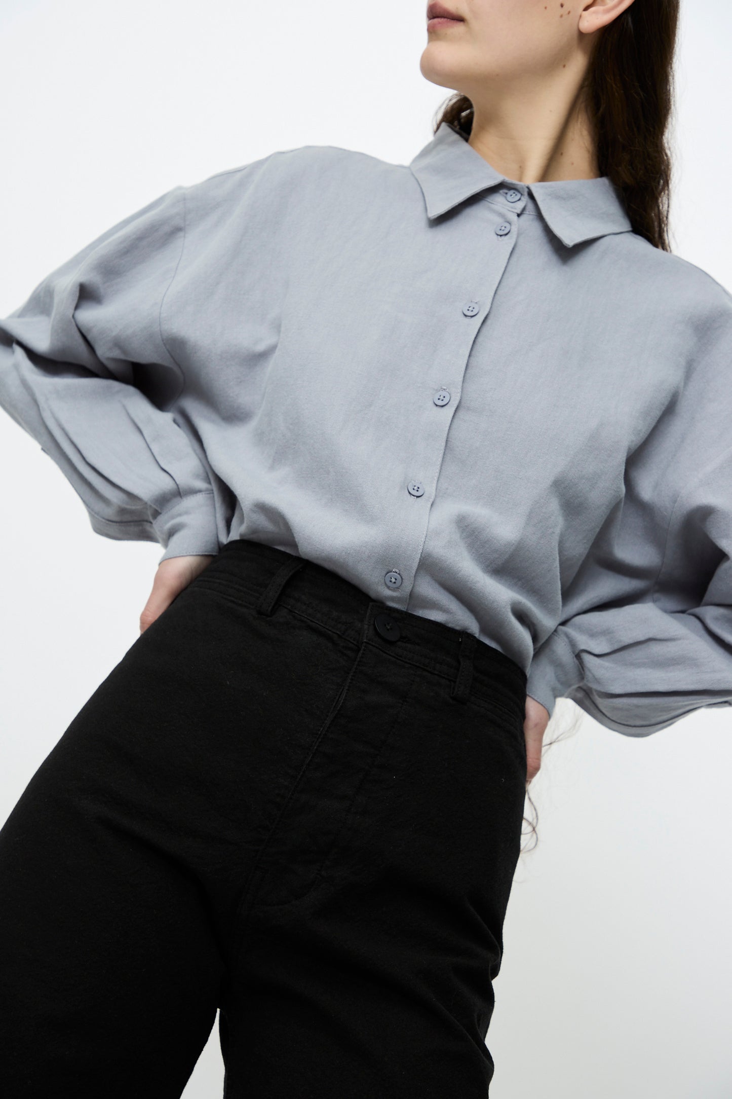 A person poses against a plain background, donning Jesse Kamm's Sailor Pant in Black with hands on hips, paired with a loose gray button-up shirt. The high-waisted pants are made from organic cotton canvas.