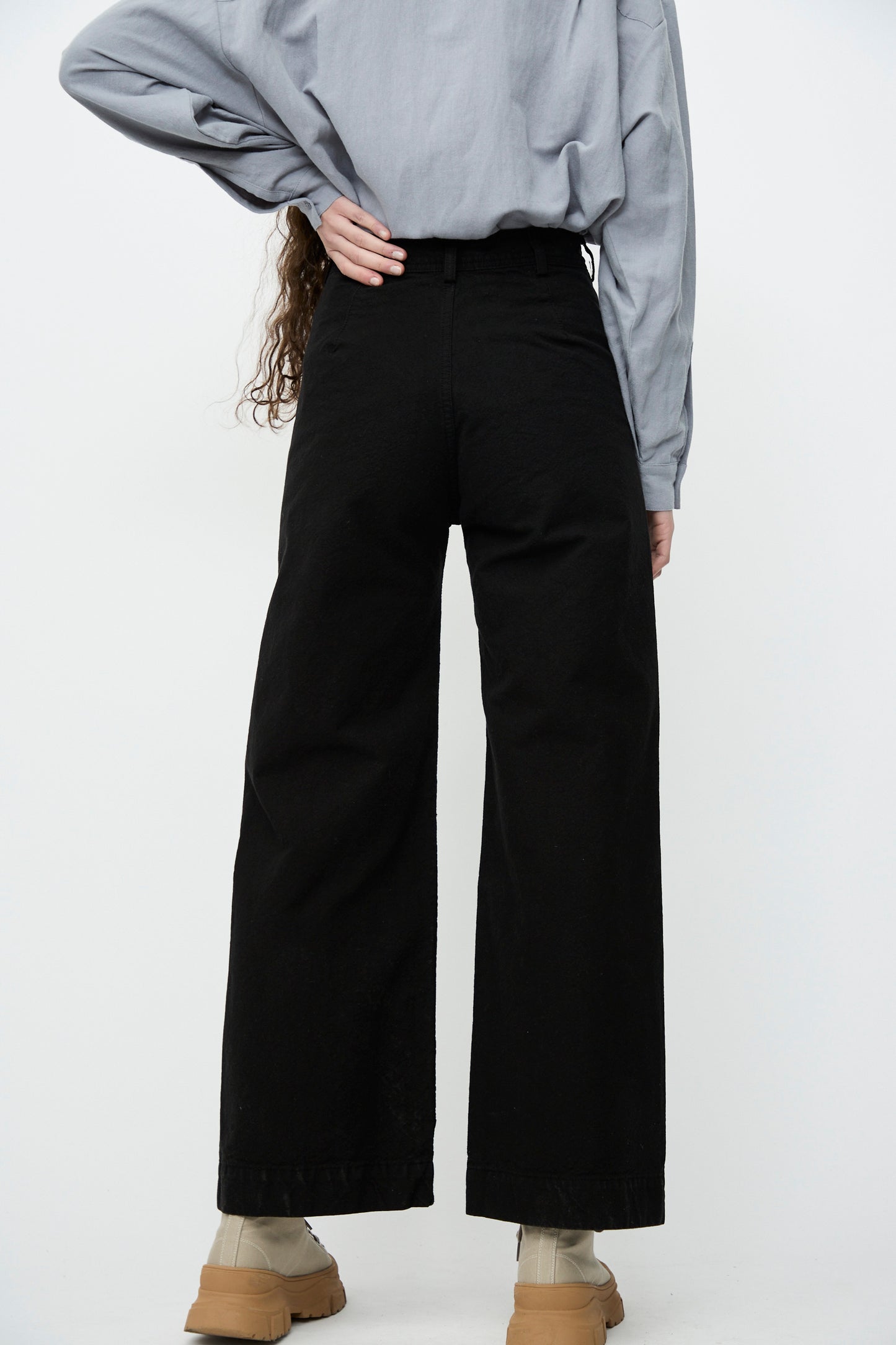 A person models the Jesse Kamm Sailor Pant in Black, made from organic cotton canvas, along with a gray shirt and tan chunky shoes, partially visible from behind.