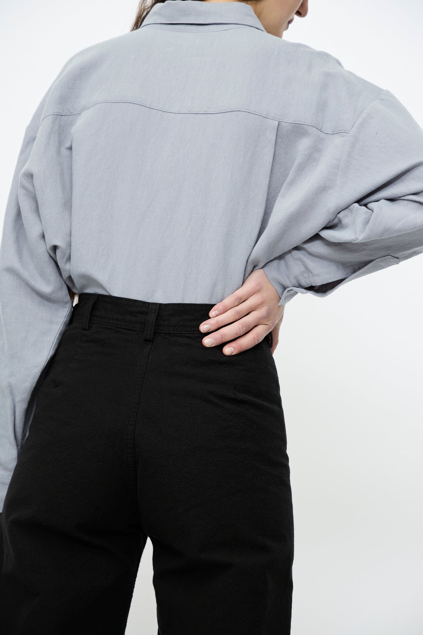 A person wearing a light gray shirt and black Jesse Kamm Sailor Pant is seen from behind with their hand on their hip against a white backdrop.