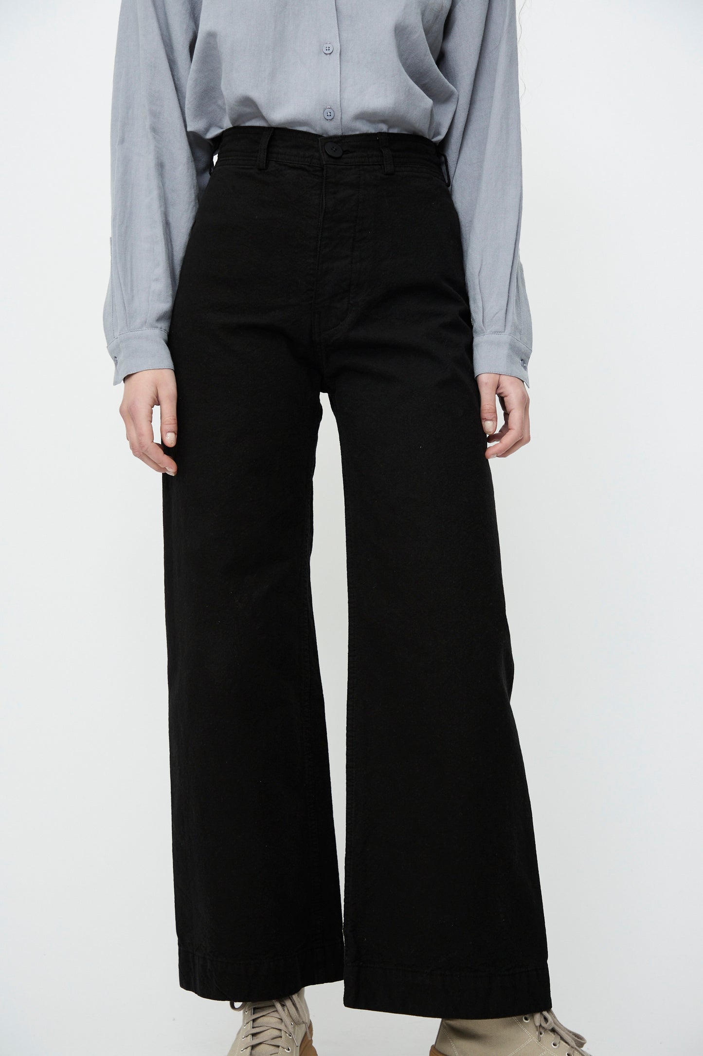 Against a white backdrop, an individual sports a light gray button-up shirt, beige shoes, and Jesse Kamm's Sailor Pant in Black, made from organic cotton canvas.