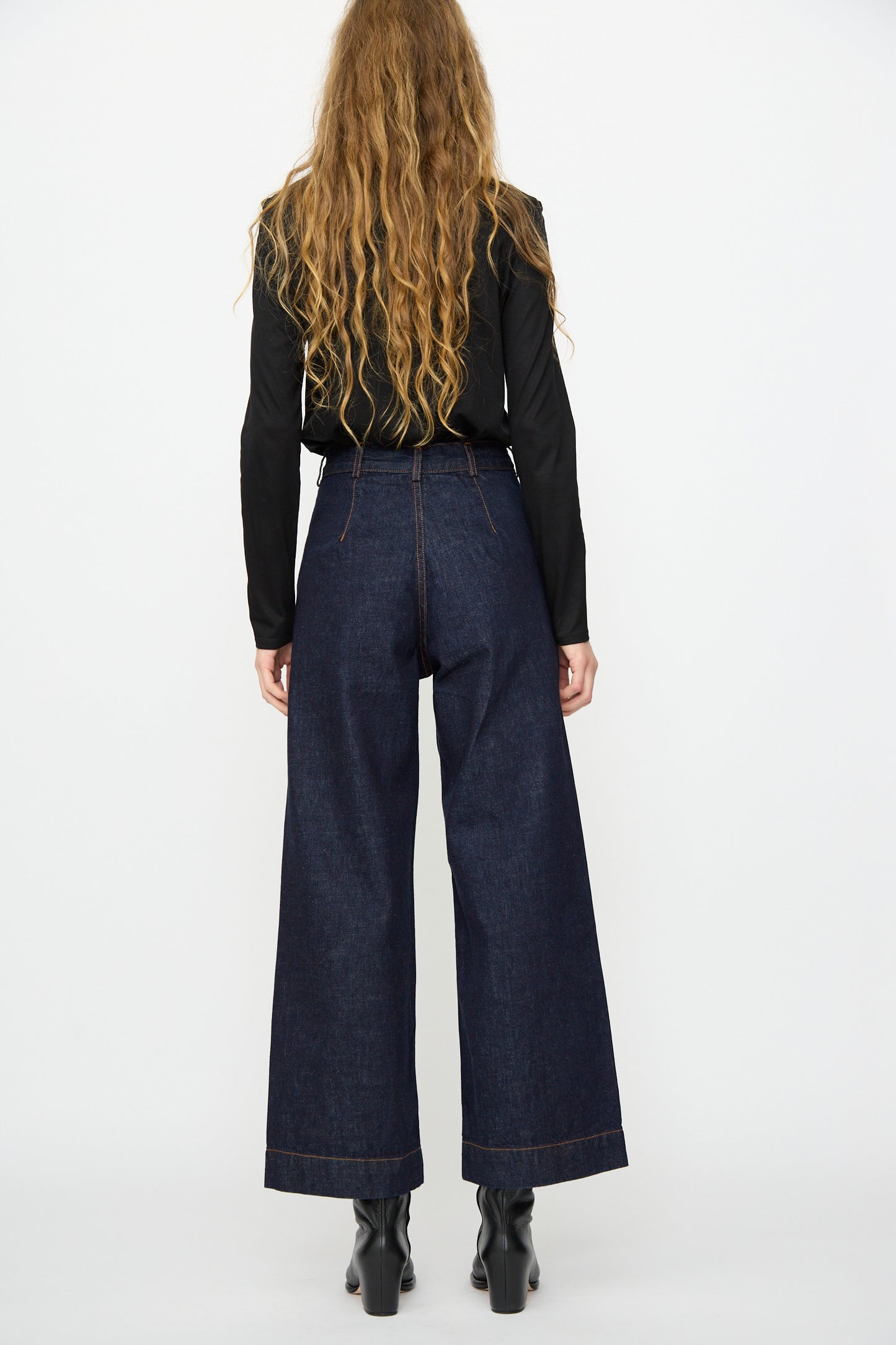 A person with long wavy hair, wearing a black top and Jesse Kamm Sailor Pant in Dark Blue, stands facing away from the camera against a plain white background.