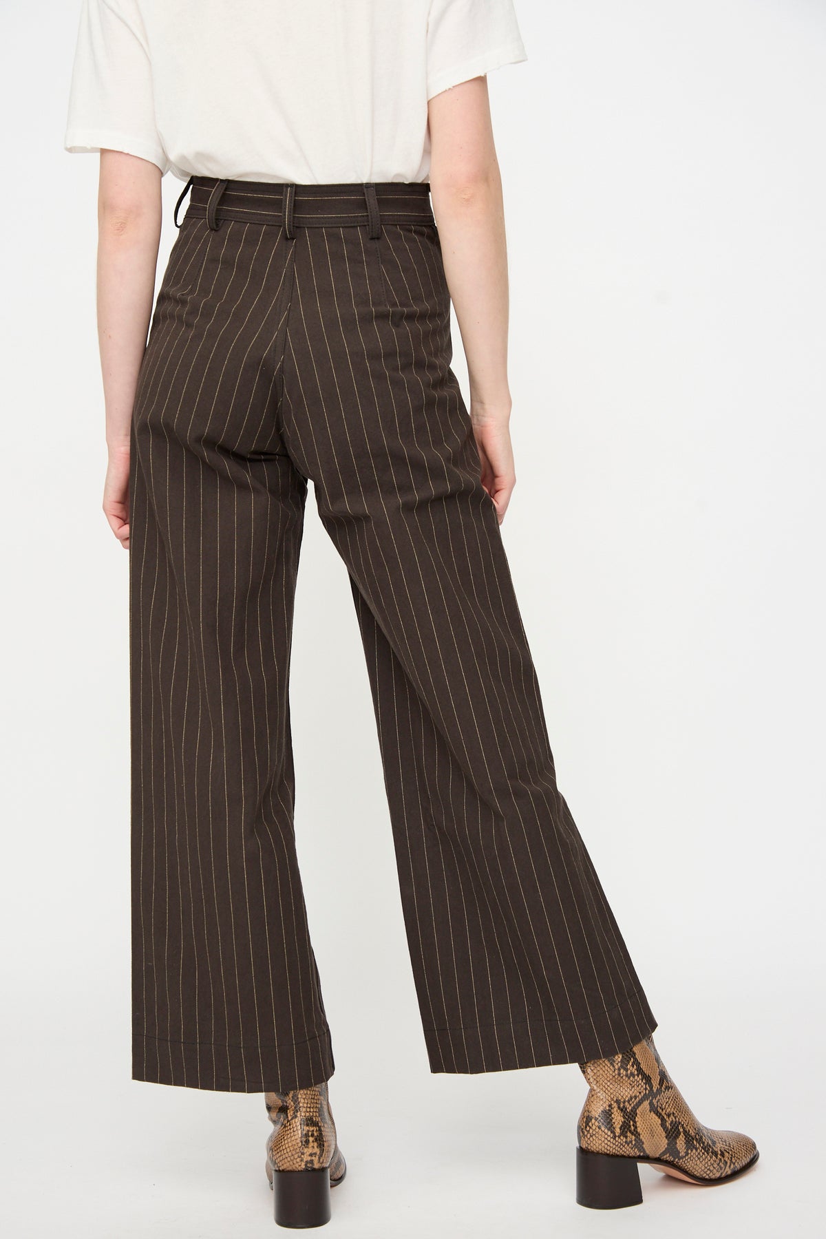 A person wearing a white shirt, Jesse Kamm Sailor Pants in Italian Stripe featuring a high waist and brown pinstripes crafted from organic cotton, paired with snakeskin patterned boots, stands with one leg slightly bent, photographed from behind against a plain background.