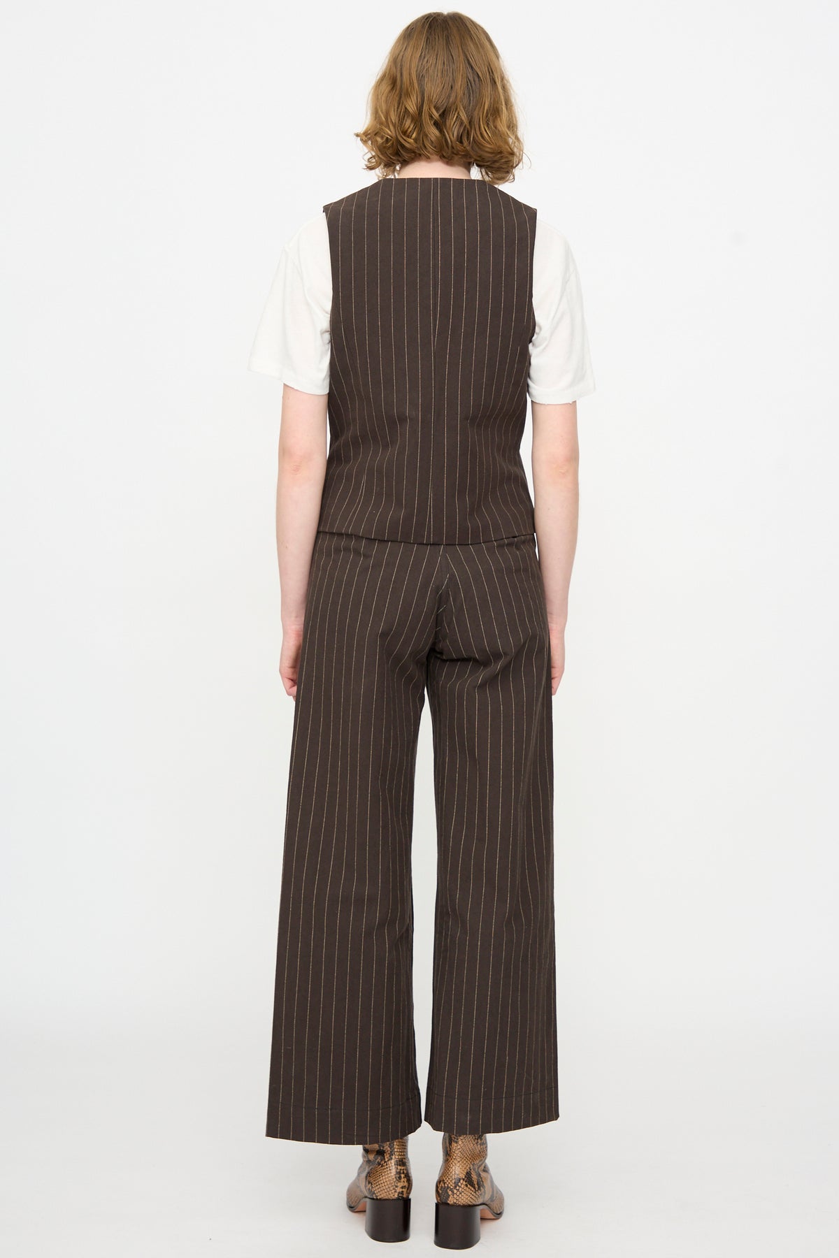 A person facing away, wearing the Jesse Kamm Trail Vest in Italian Stripe—a brown pinstripe waistcoat with matching striped cotton pants—over a white short-sleeve shirt, stands against a plain white background.