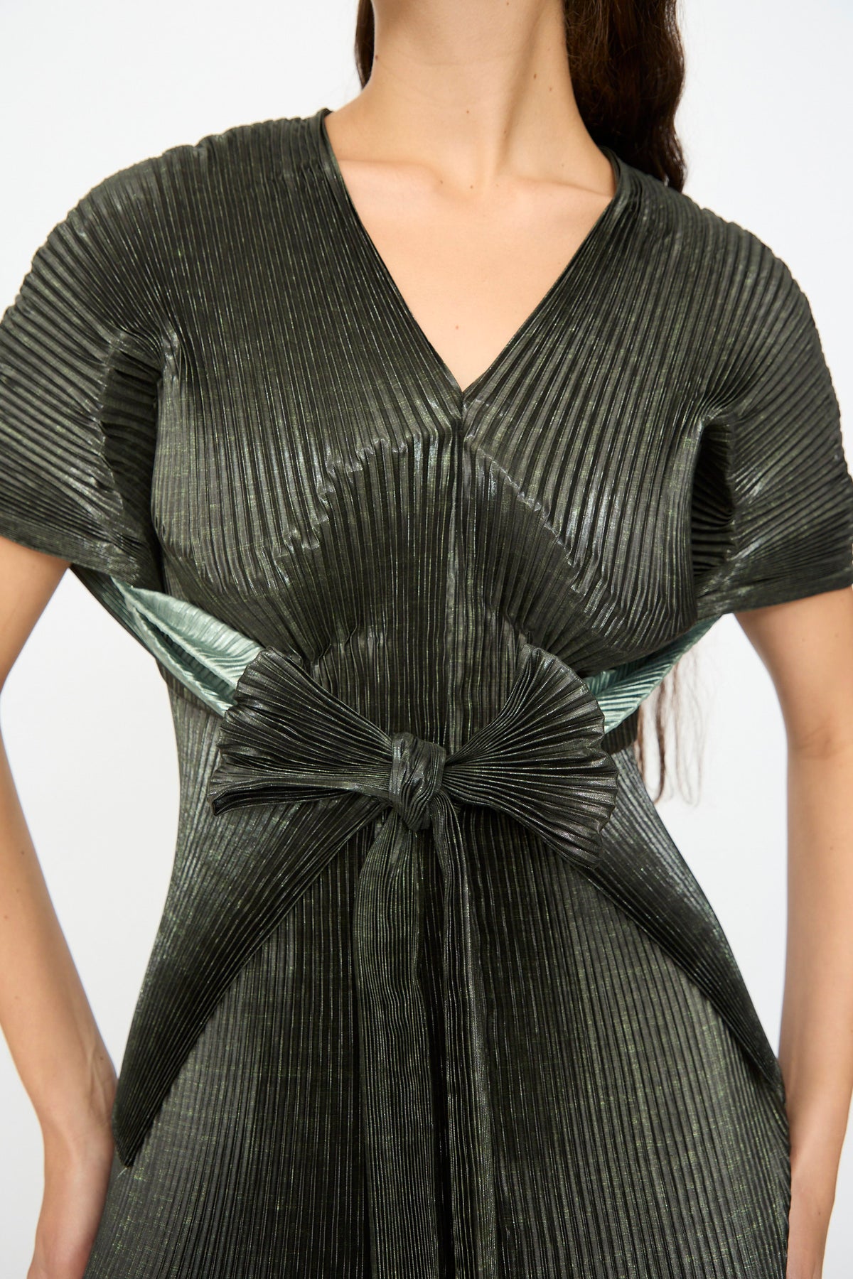 A woman wears the Edgar Pleated Poncho in Moss Metallic by Julia Heuer, featuring a dark, pleated design with a V-neck, short sleeves, and a large matching bow tied at the waist.